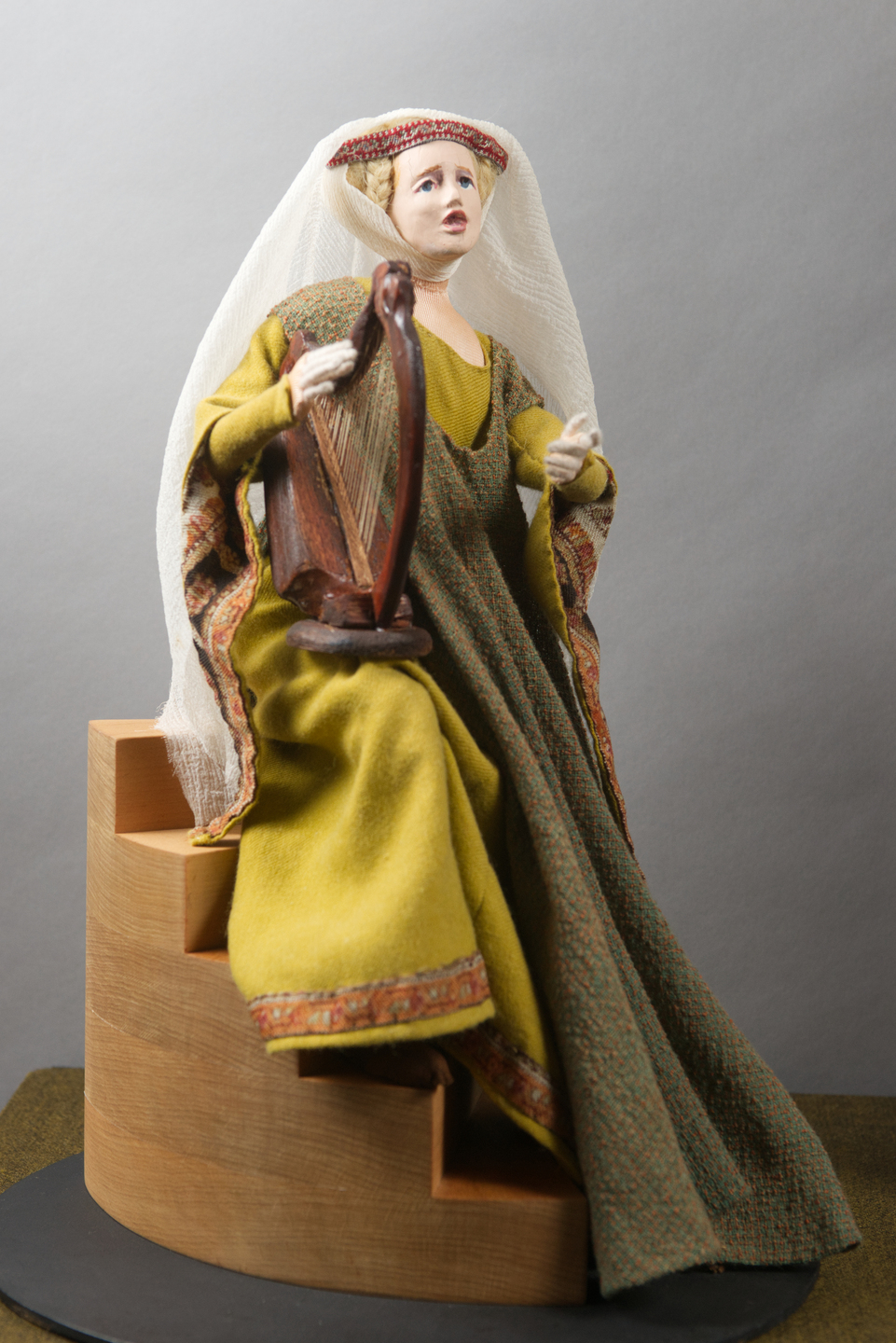 A model of a medieval woman seated on wooden steps, dressed in a long green gown with patterned sleeves and a veil. She holds a small harp in her lap, her posture suggesting she is preparing to play.