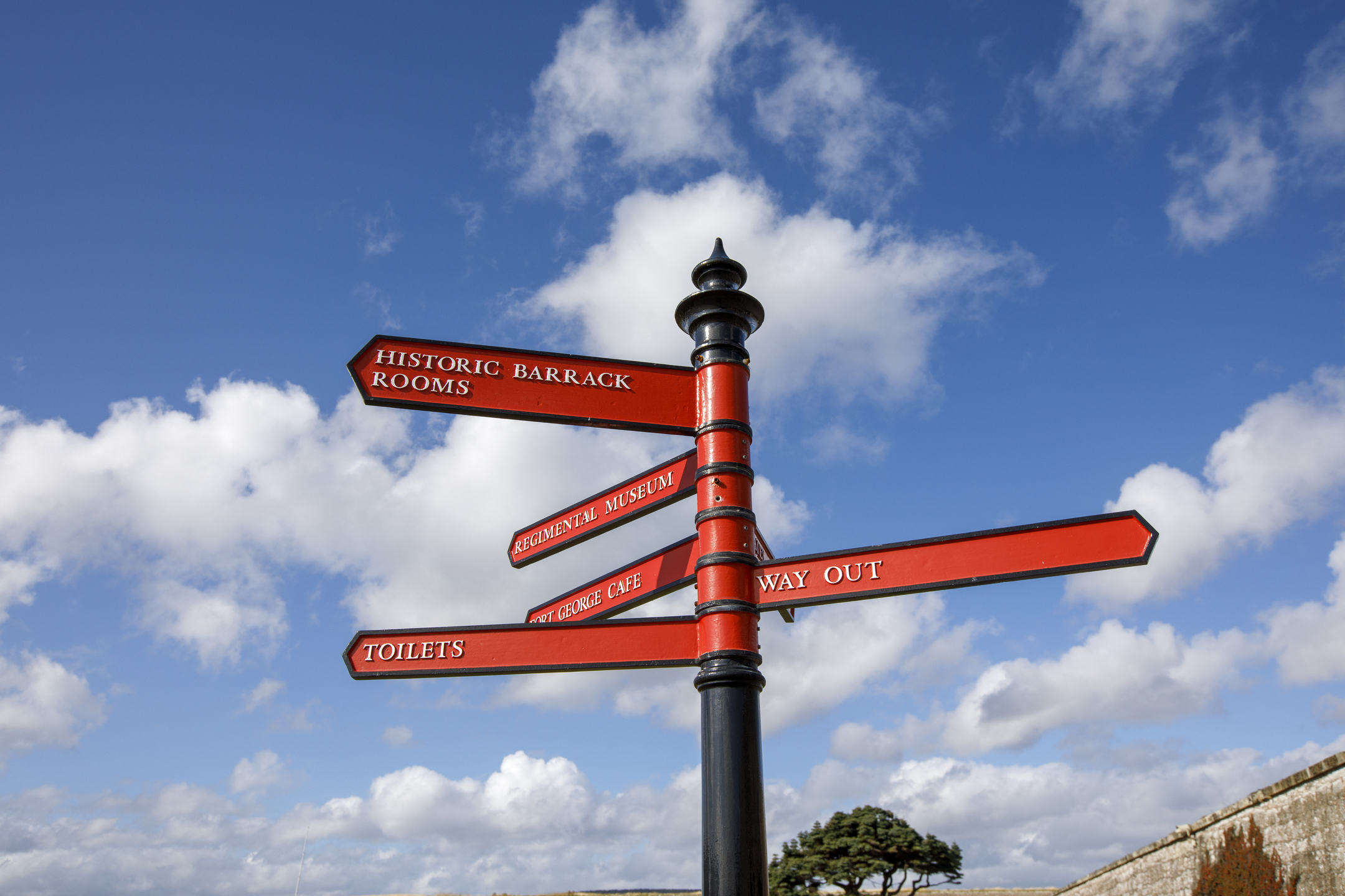 A bright red signpost at Fort George pointing toward visitor areas including the historic barrack rooms, museum, café, toilets, and the way out, set against a blue sky with scattered clouds.
