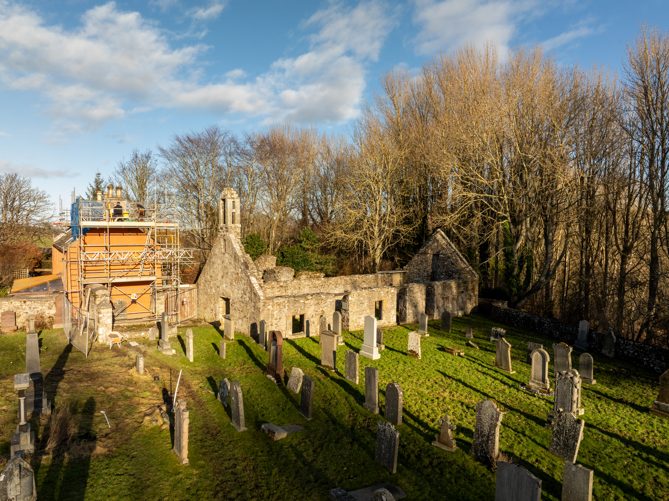 View of ruined and roofless stone church surrounded by a grassy graveyard and bare trees under a blue sky with clouds. Scaffolding work on another building can be seen to the left. 