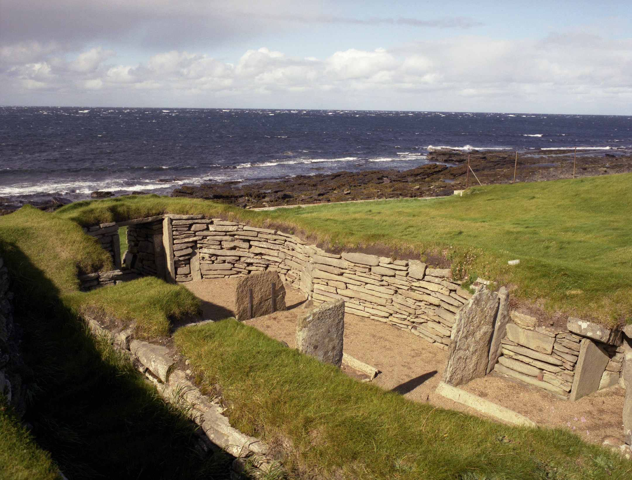 The stone structure of the Knap of Howar sits on grassy ground above a rocky shoreline, with waves breaking along the coast.