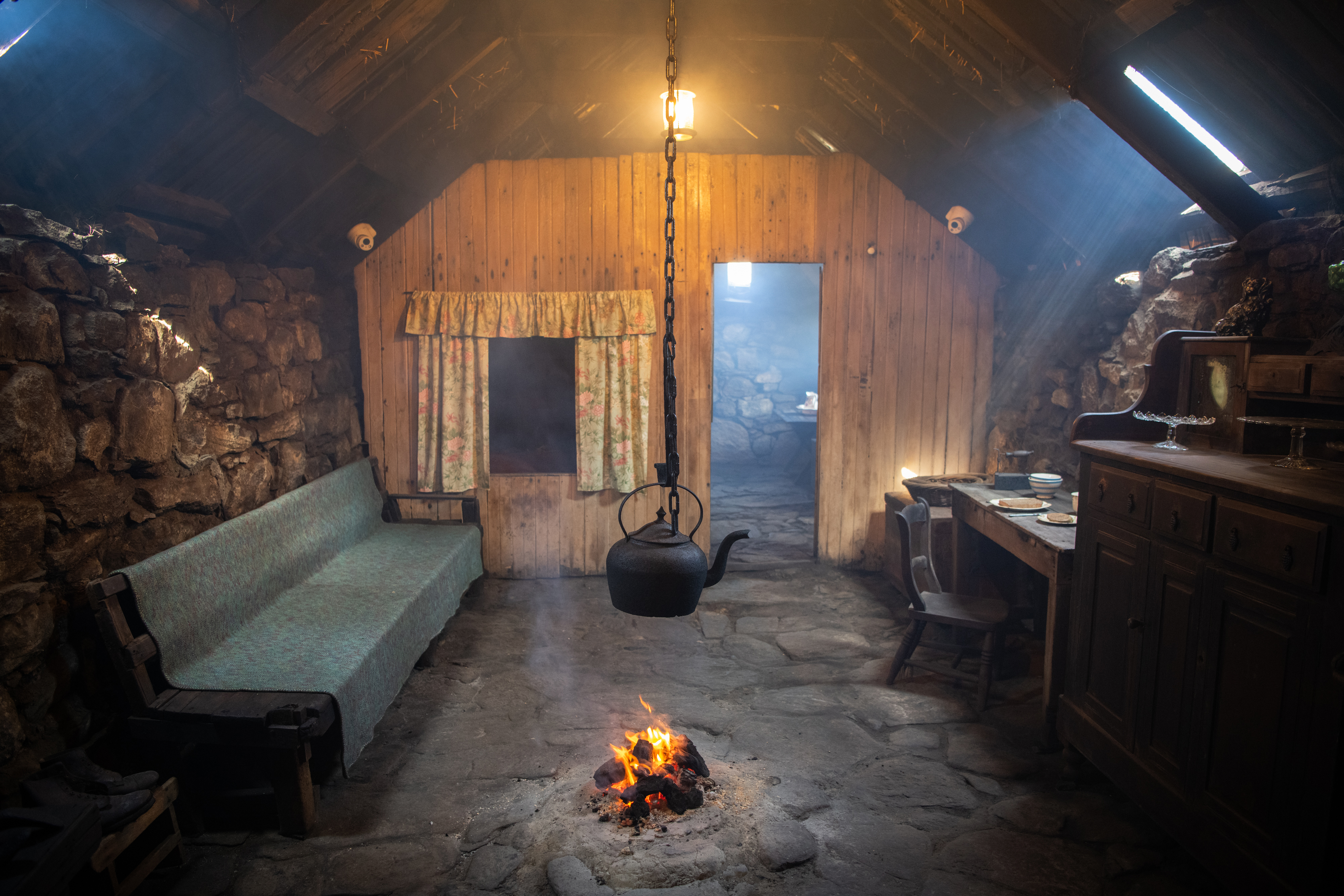 A living space in a stone and wood walled room, with a stone floor. There is a long bench and other furniture, and in the middle of the room an iron kettle hangs on a chain above a small open fire 