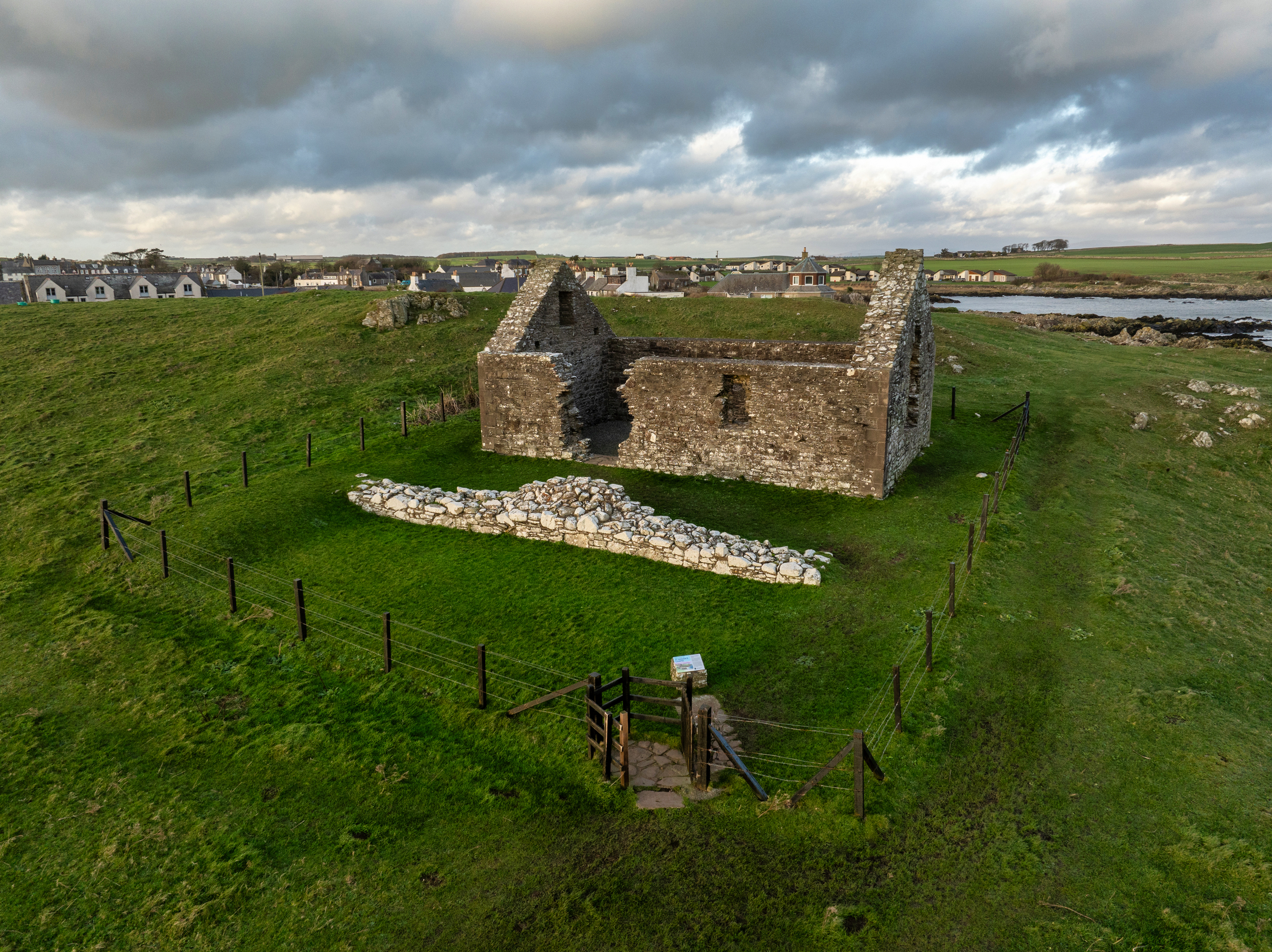 Aerial view of St Ninian’s Chapel ruin within a fenced grassy enclosure, with a village and open fields in the background under a cloudy sky.