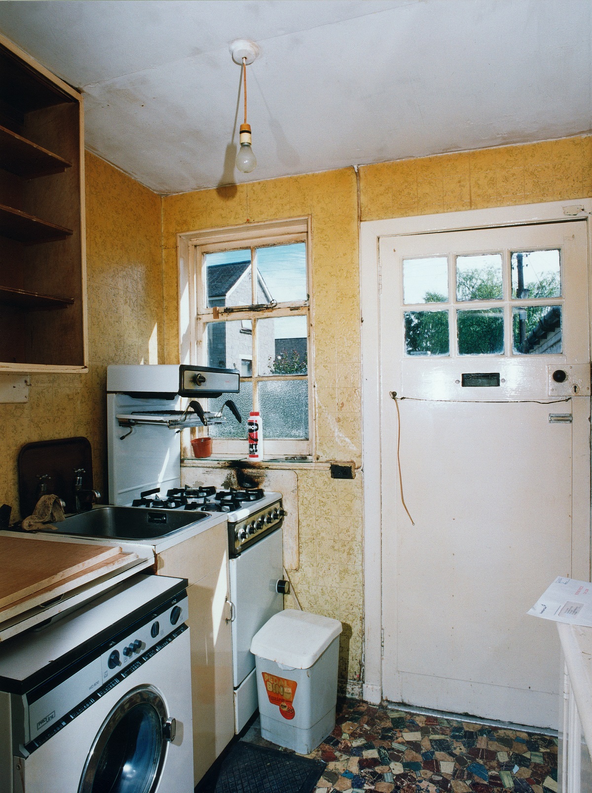 Small, dated kitchen at Sunnybrae Cottage with yellowed walls and a patterned linoleum floor. The space includes a white gas stove with an overhead extractor, a stainless steel sink, and a washing machine in the foreground. There is a single window above the sink. A bare lightbulb hangs from the ceiling. 