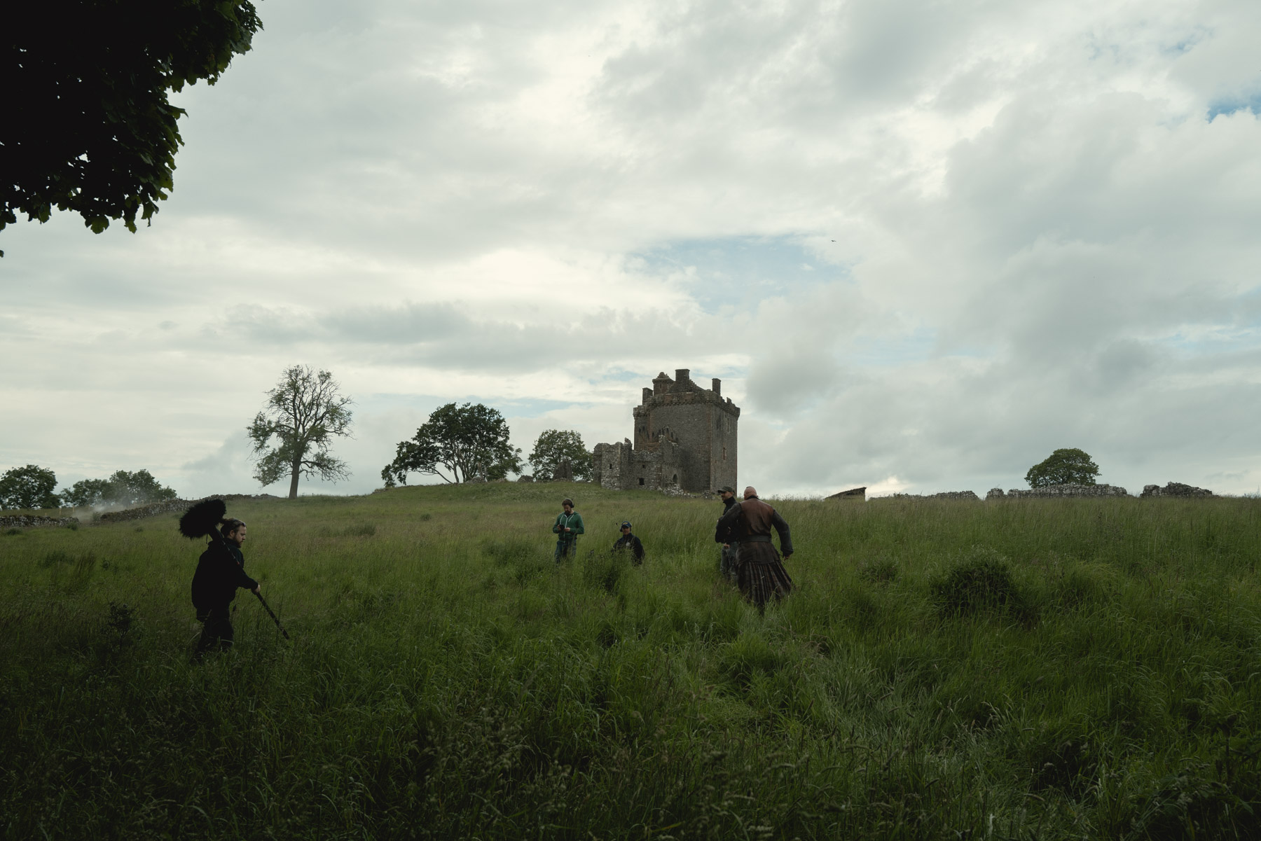 Behind the scenes at the filming of a historical drama. Cameras film an actor in a kilt walking through long grass towards an imposing hilltop castle.