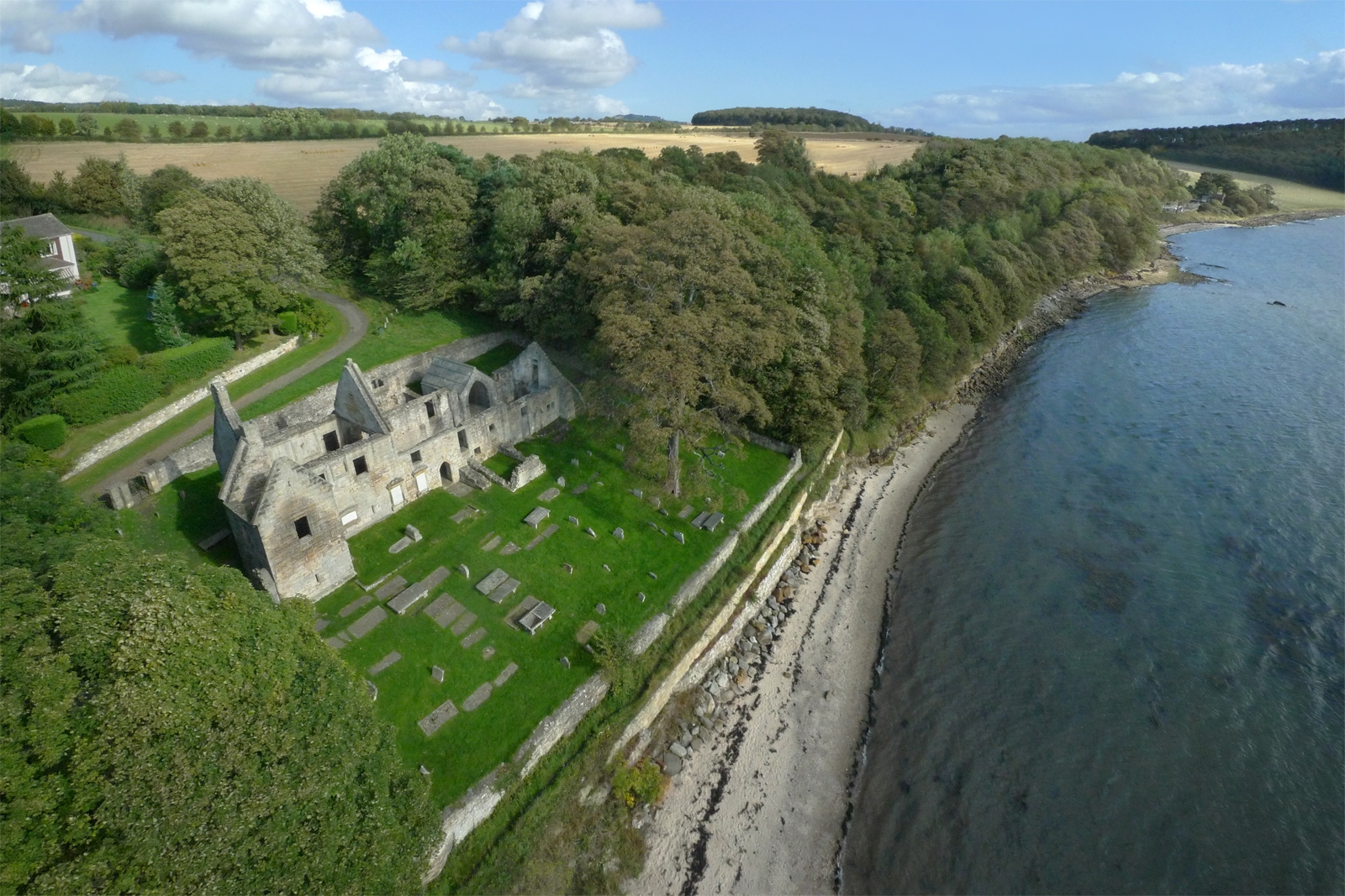 Aerial view of the roofless stone ruins of St Bridget’s Kirk and its graveyard, set on a grassy terrace beside a wooded hillside and a curving shoreline.