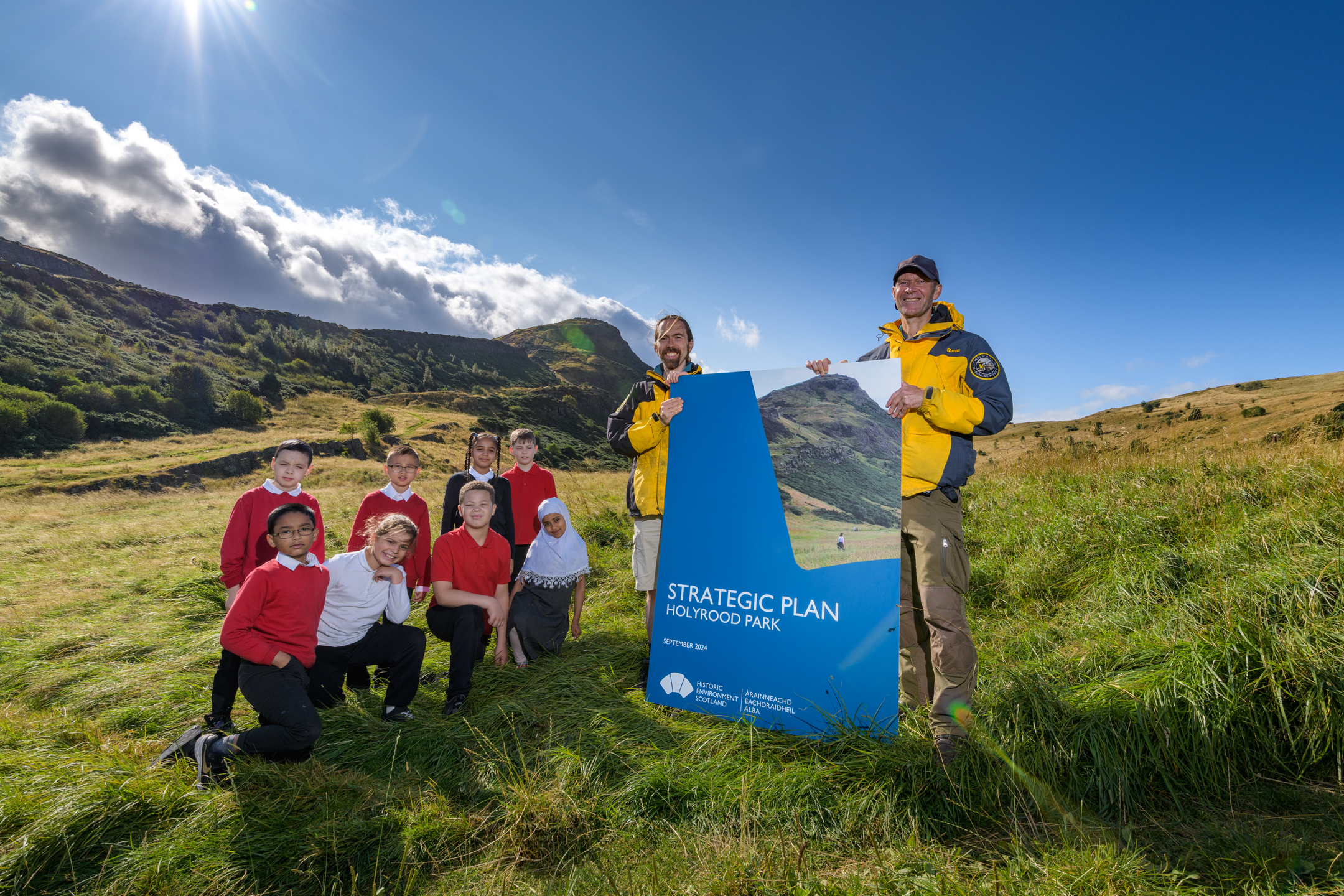 Group of adults and children standing on a grassy hillside in Holyrood Park beside a large blue sign reading “Strategic Plan – Holyrood Park,” with hills and blue sky in the background.