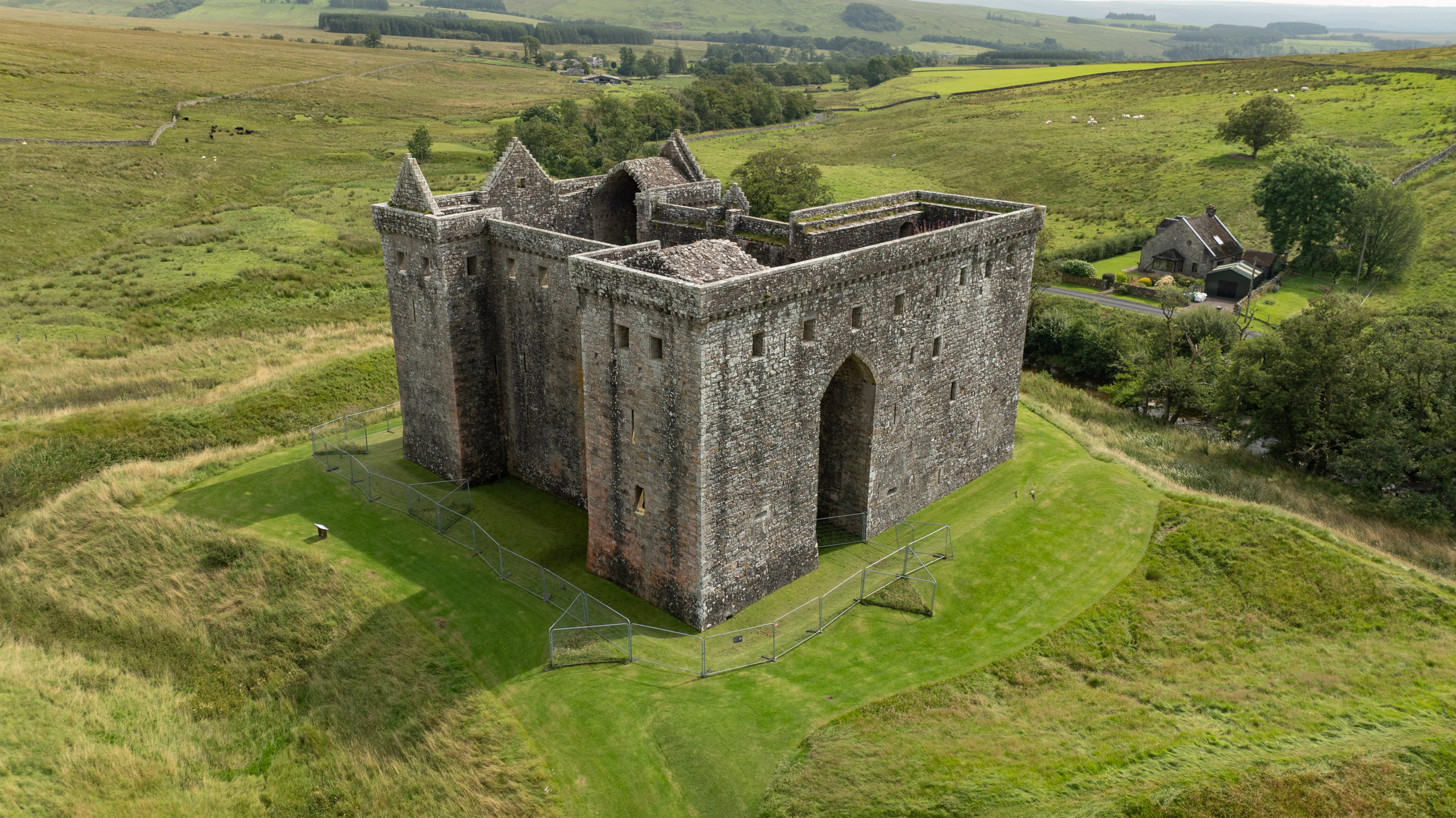 Aerial view of Hermitage Castle surrounded by grassy landscape and distant hills.