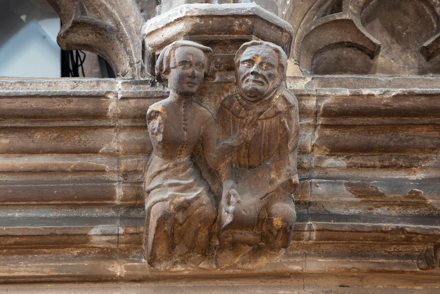 Stone carving of two seated robed figures, likely a husband and wife, one gesturing with a raised arm, on a ledge of the cathedral.