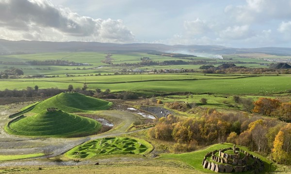 A green landscape on a cloudy day. To the right, there is a small circular rock structure, to the left you can see two unusual looking mounds. Behind them you can see trees and fields.