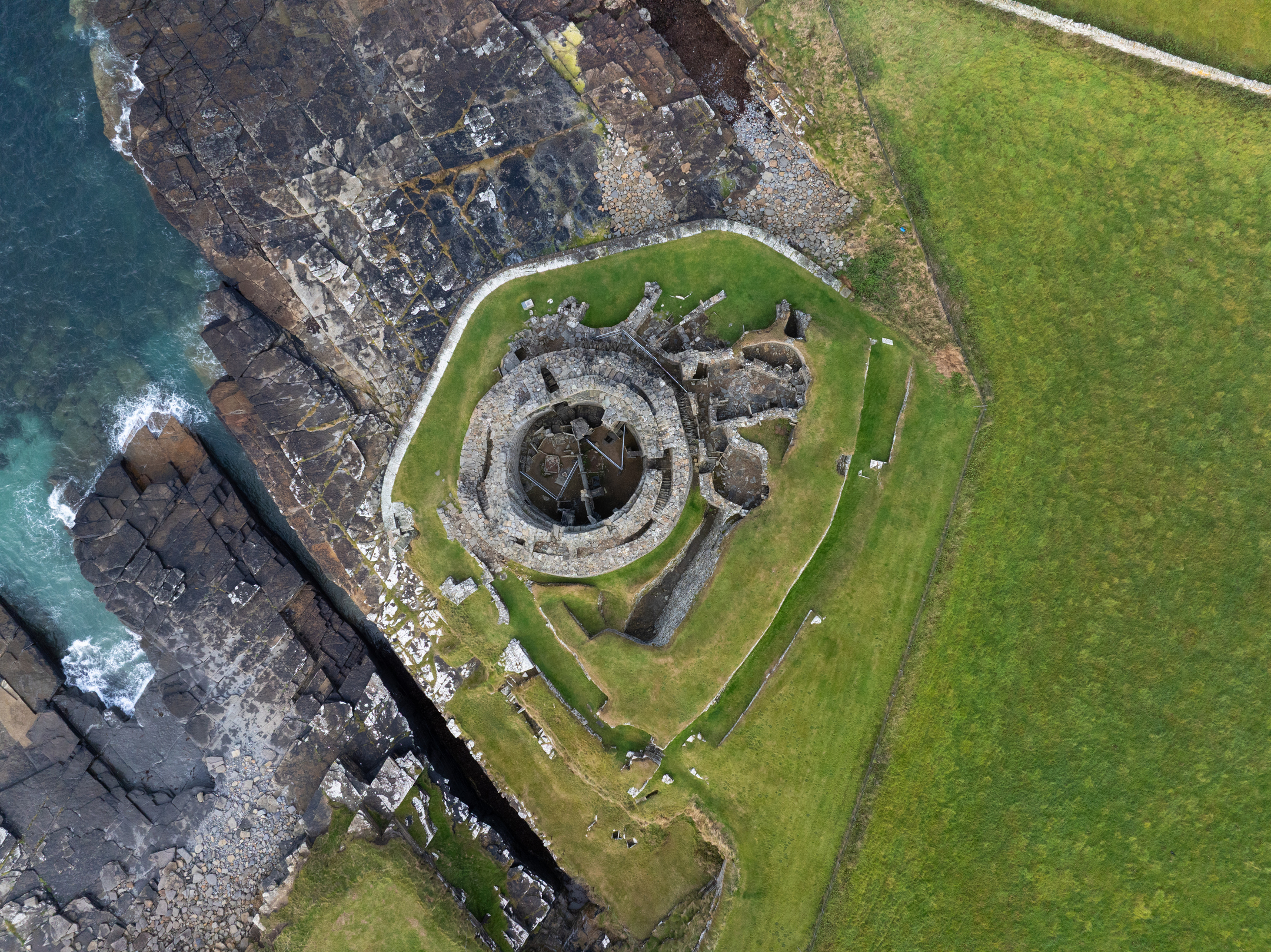 Top-down aerial image of Midhowe Broch revealing the full ring-shaped structure, central chamber, and surrounding grassy landscape meeting the rocky coastline.