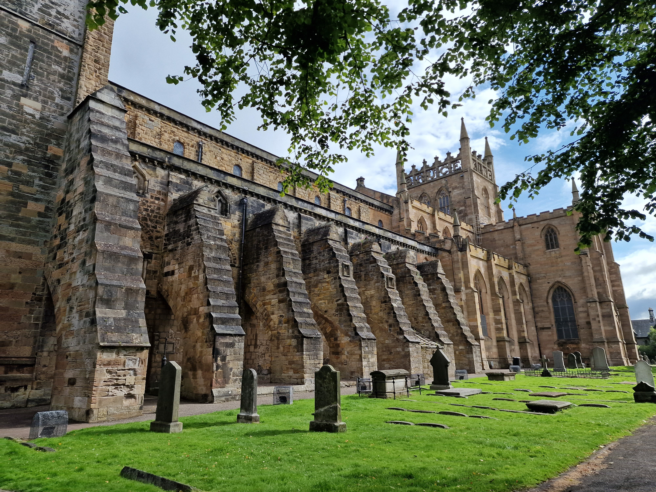 A row of heavy stone buttresses supports the side wall of a medieval abbey, with gravestones and a green churchyard lawn in the foreground.