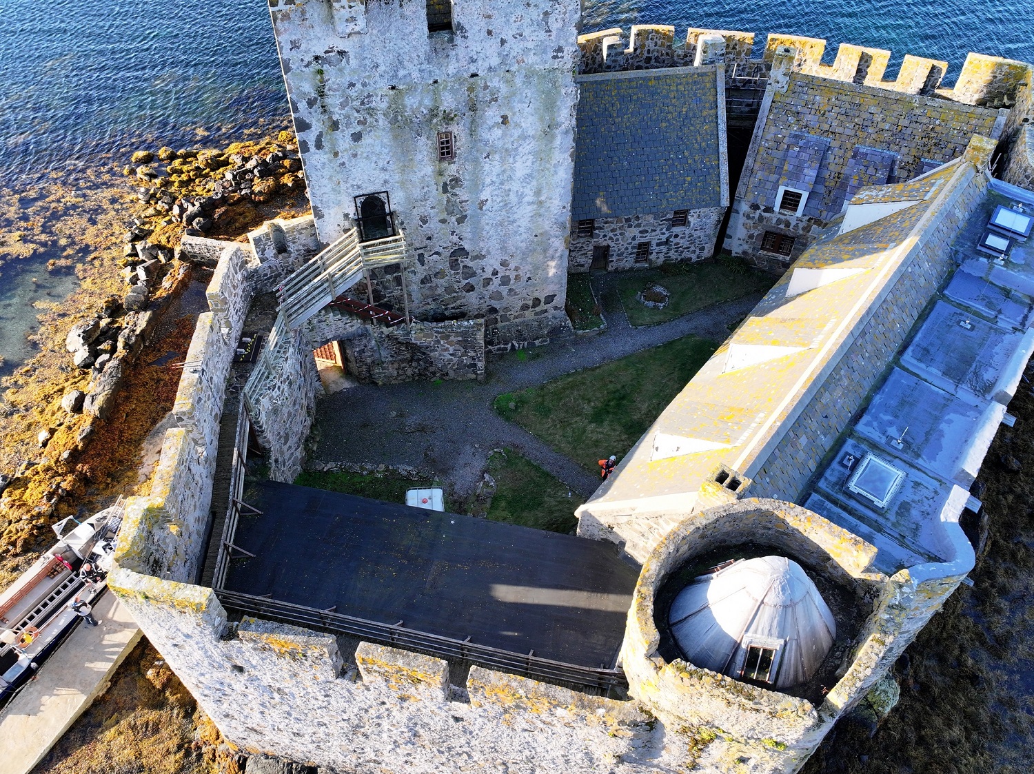 Aerial view looking down into a stone castle’s interior courtyard and walls, showing walkways, roofs, and the surrounding shoreline.