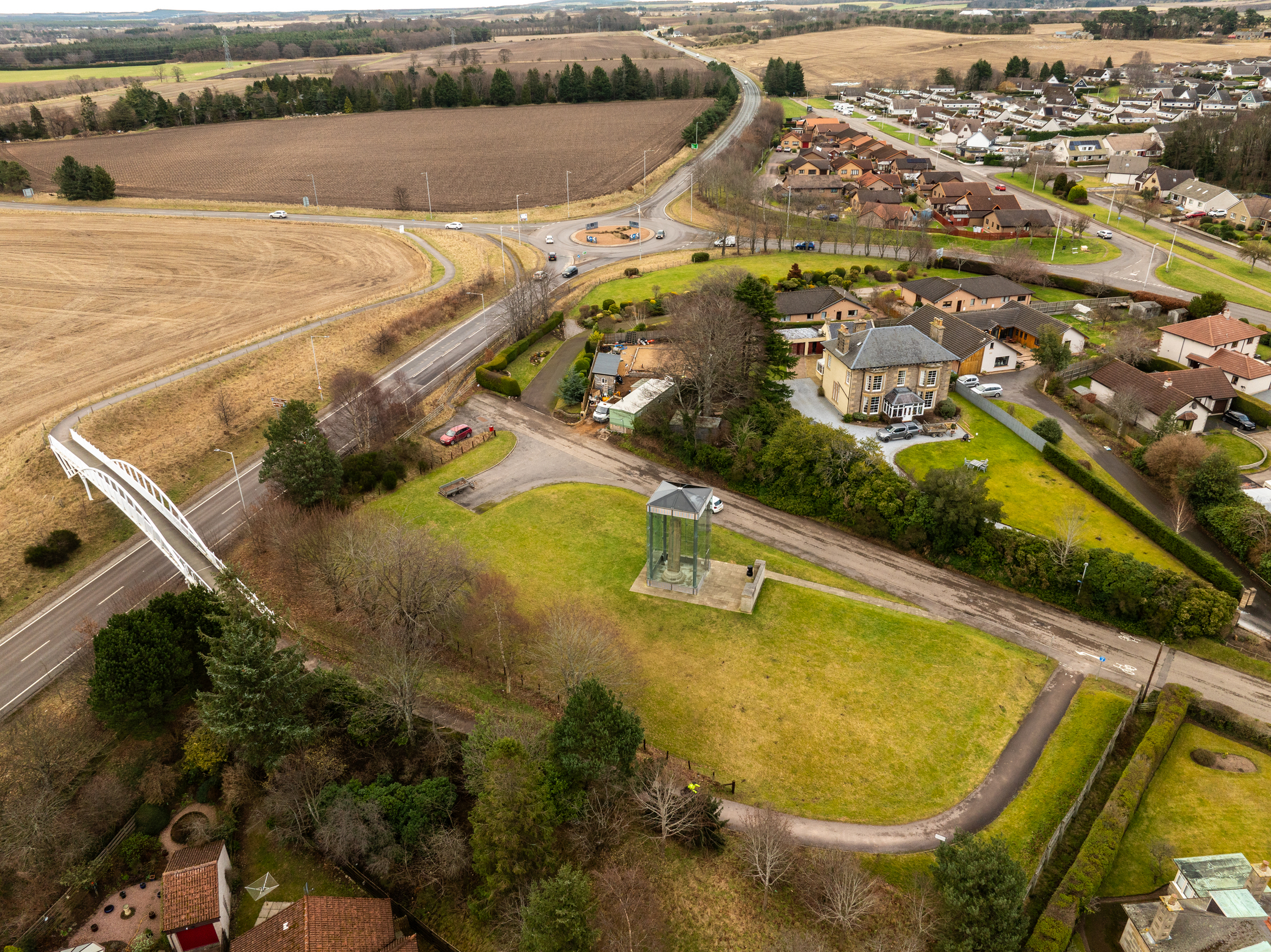 Wide aerial view of Sueno’s Stone site showing its glass enclosure on a grassy plot near a curved road. The surrounding landscape includes agricultural fields, residential houses, and a network of roads under an overcast sky.