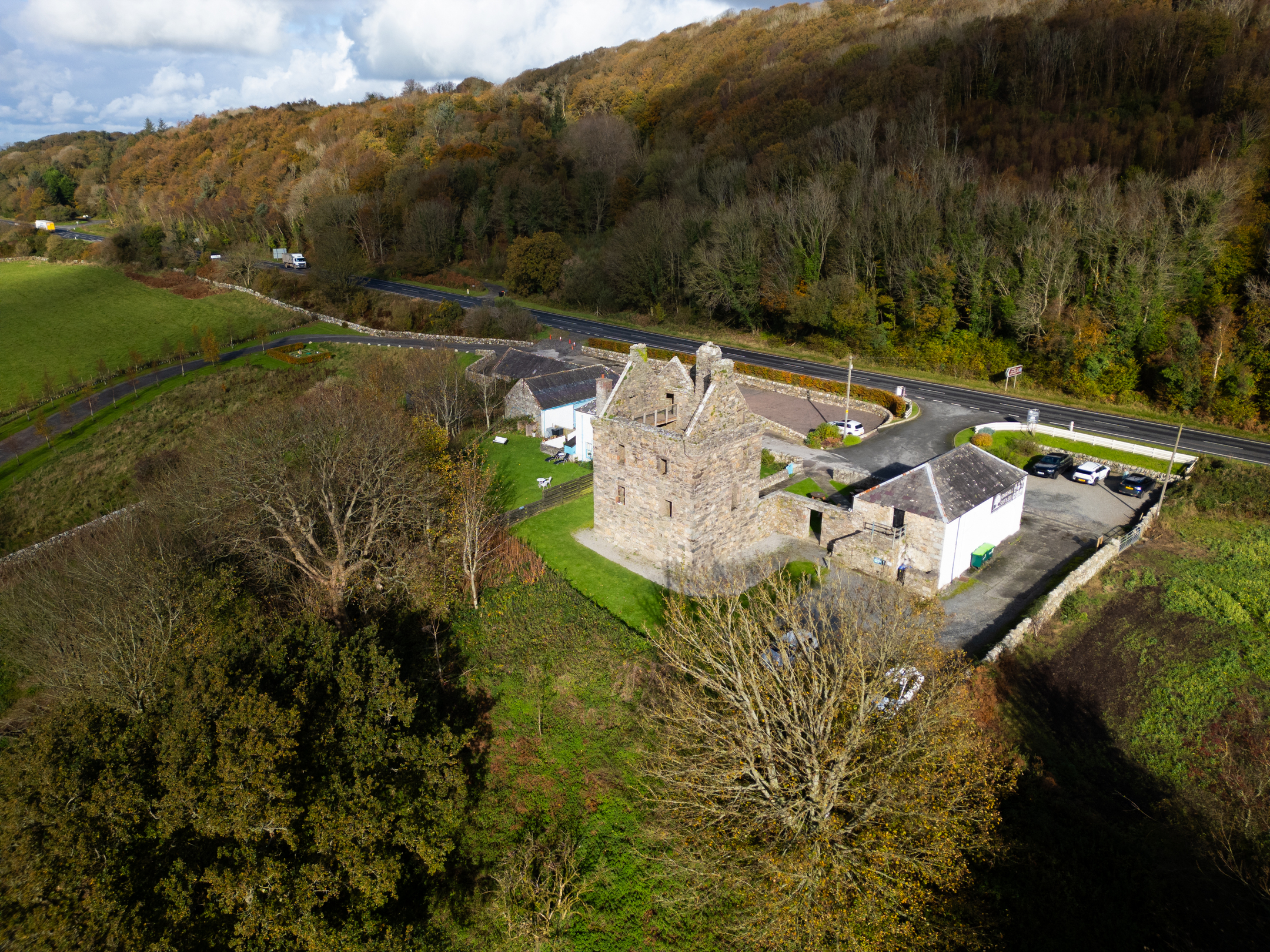 A drone‑style shot of Carsluith Castle and its surrounding buildings, set beside a road and backed by wooded hills in autumn colours.