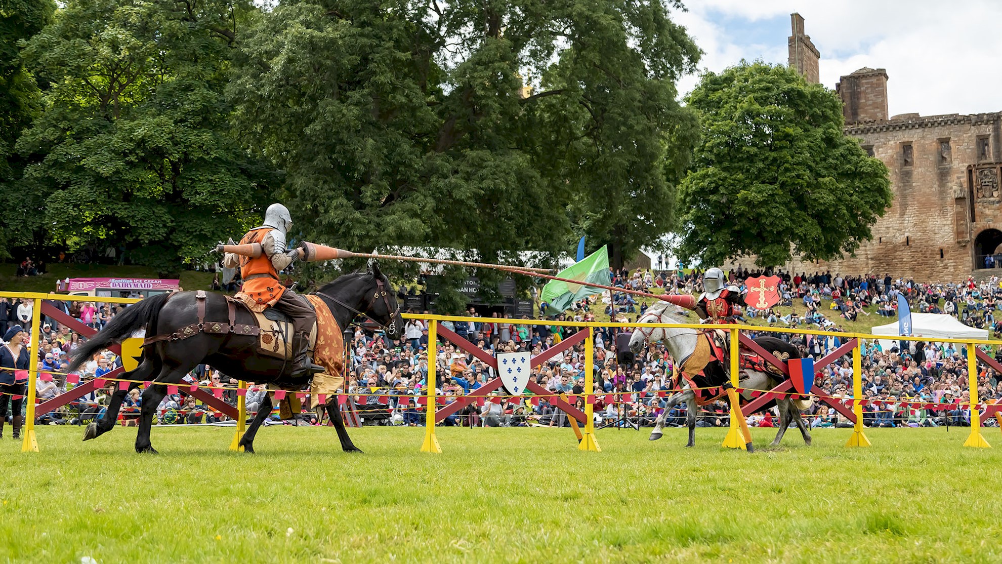 Two knights on horseback charge at each other with lances in hand, in front of a crowd of people. Linlithgow palace sits in the background. 