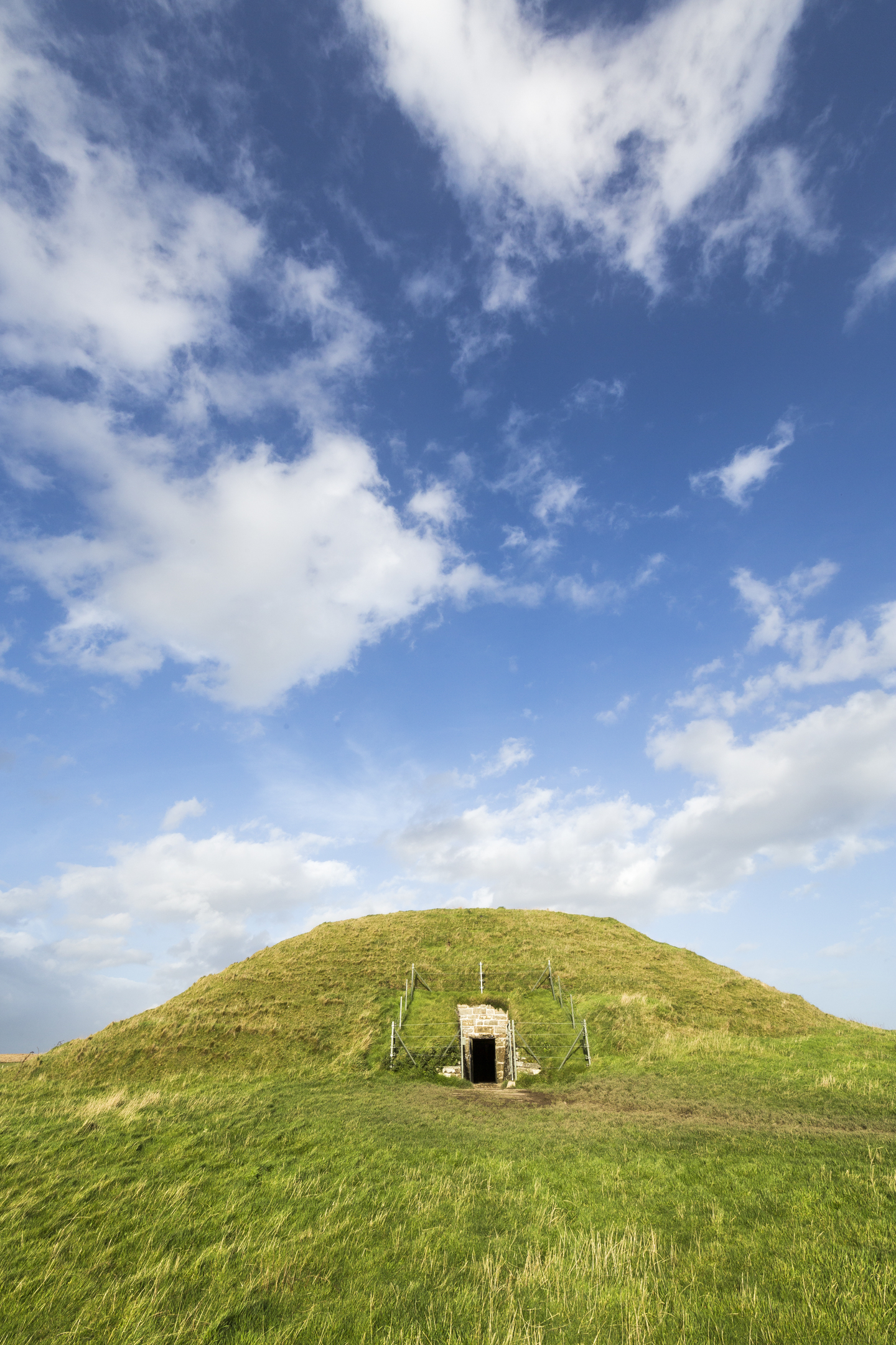 Grass-covered mound with entrance under a blue sky with clouds.