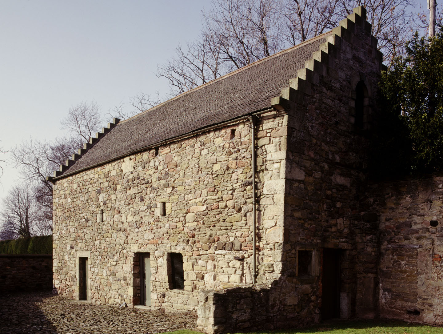 Long stone building with two doors and windows, cobbled yard in foreground and a slate roof.