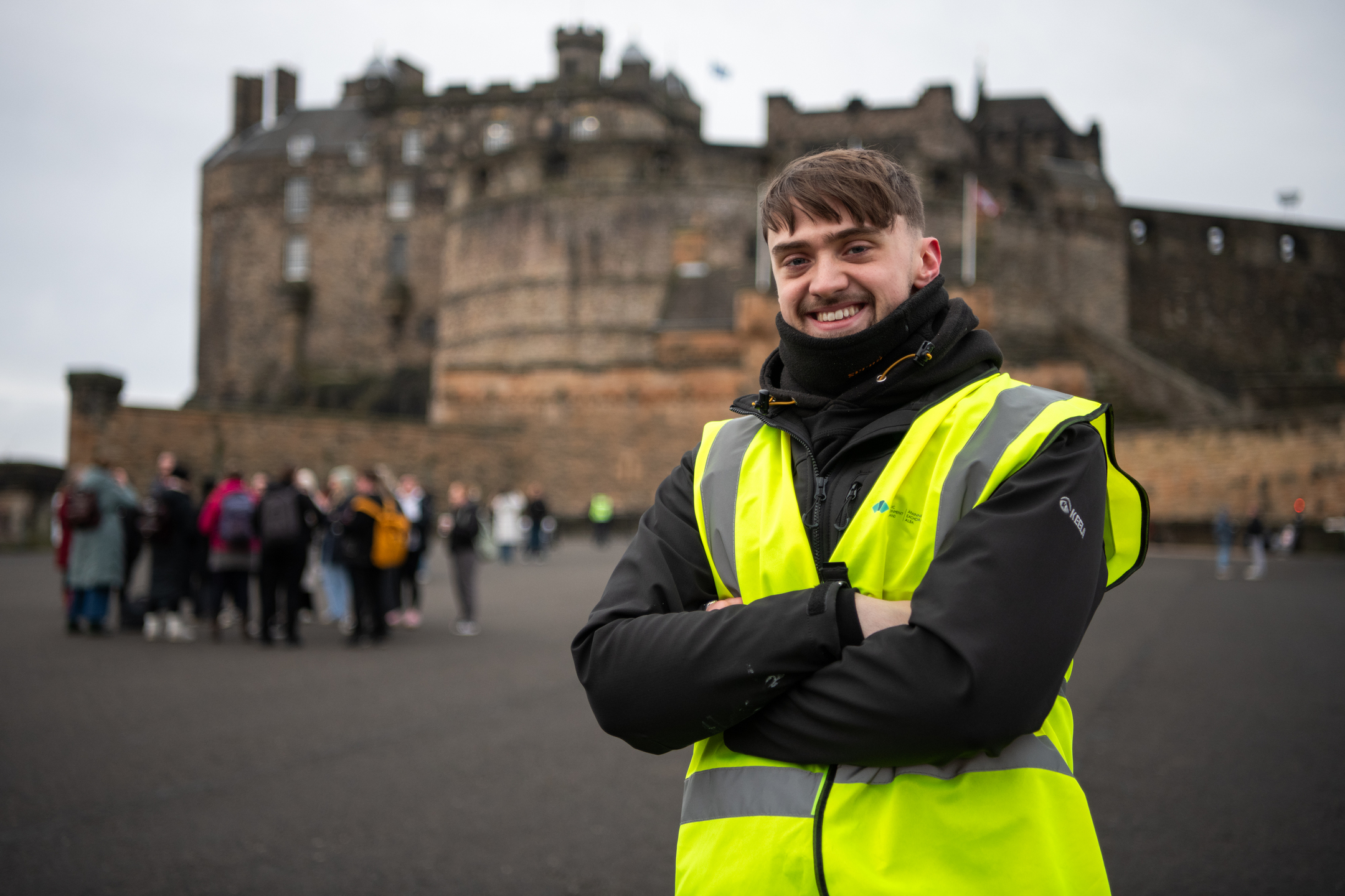 A person wearing a high-visibility yellow vest and dark outdoor clothing stands with folded arms in the foreground of Edinburgh Castle.