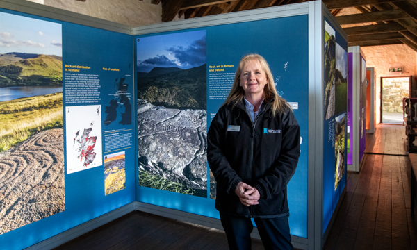 A woman with long blonde hair wearing a black fleece with the Historic Environment Scotland logo on it stands facing the camera in a room with dark wooden floorboard and wooden beams across the ceiling above. Behind her are display boards covered in text and images of rock art across Scotland.