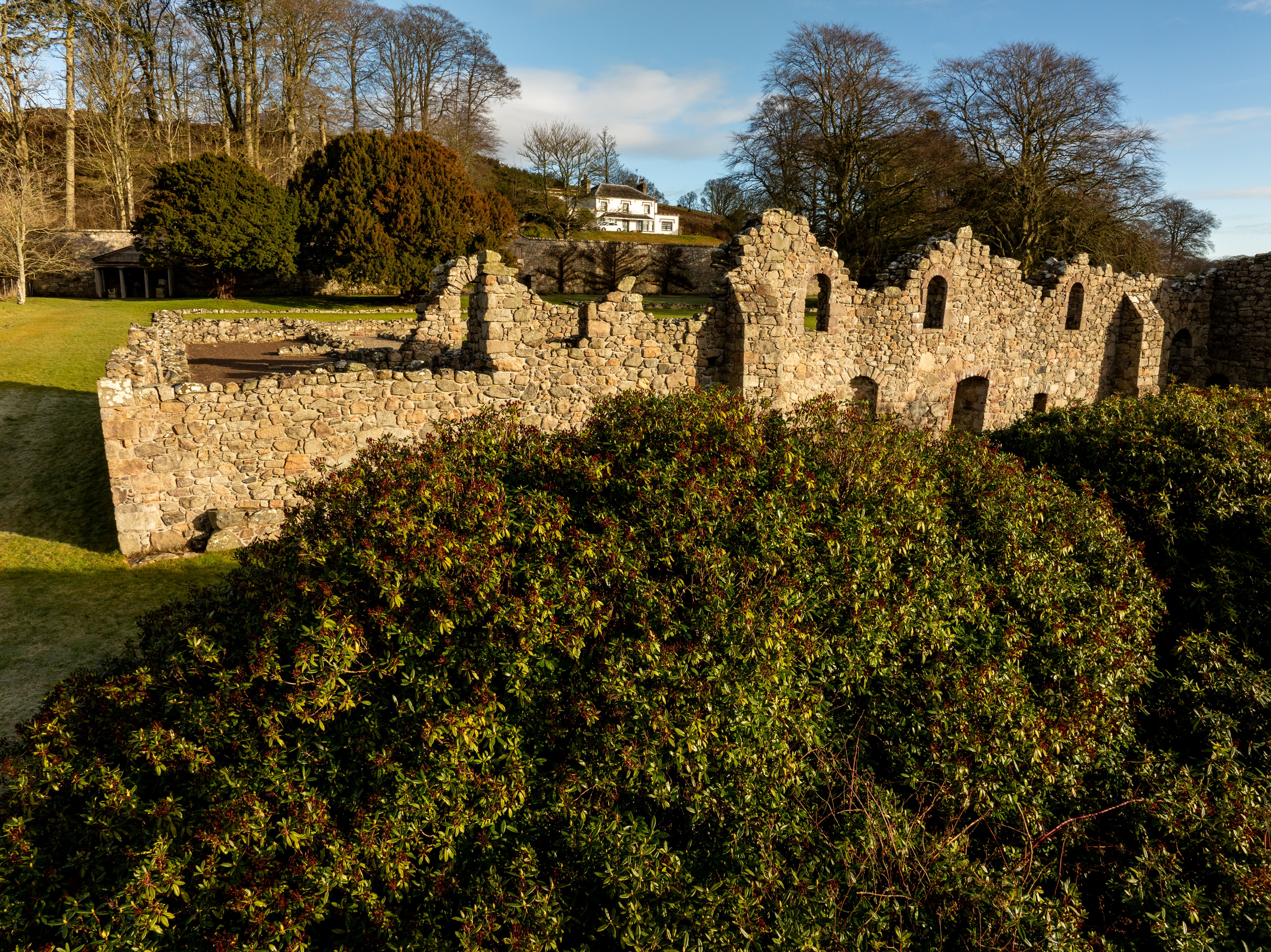 View of ruined wall and greenery. Trees and a white building can be seen in the background under a blue sky.