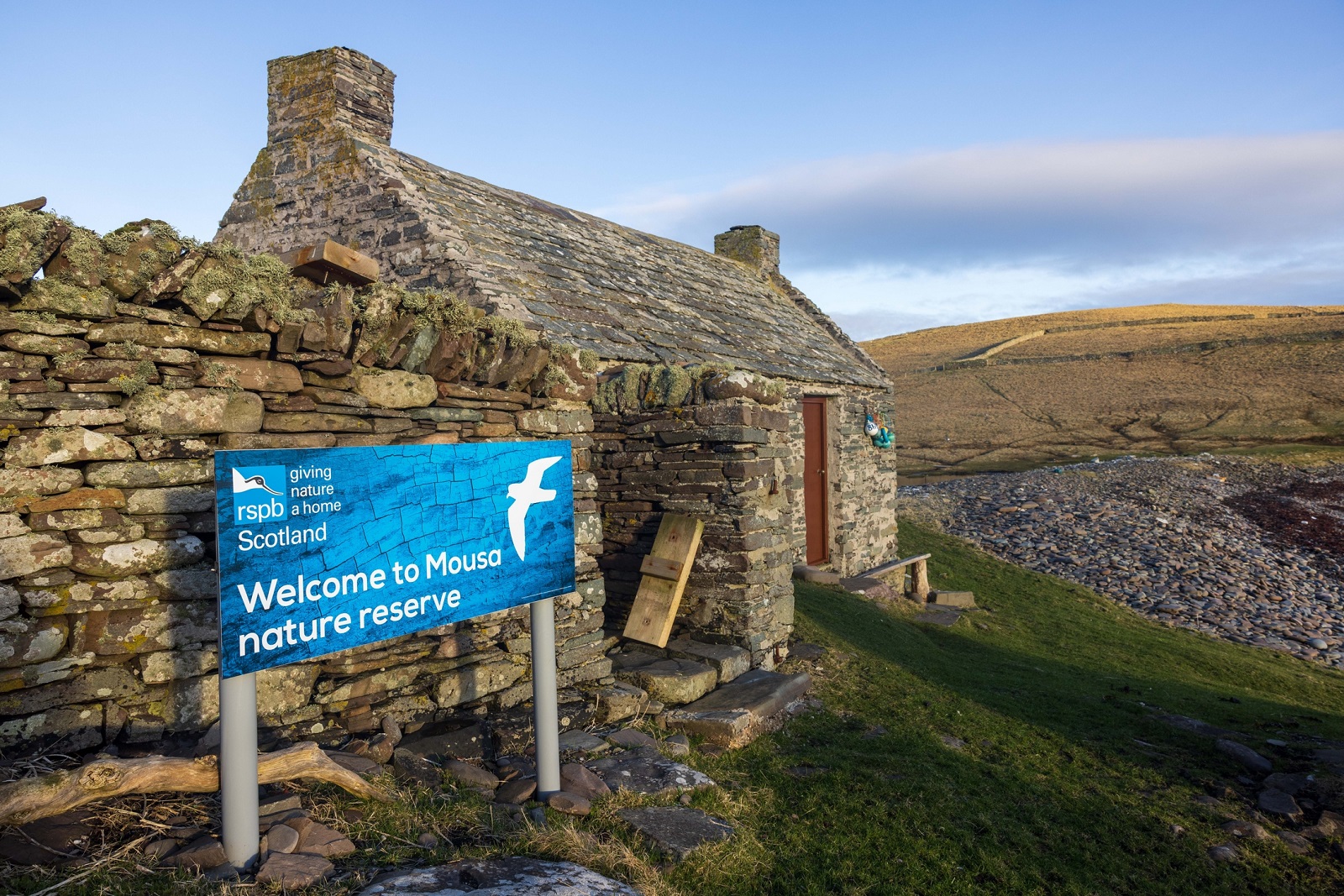 small drystone cottage with a large blue sign outside with reads "Welcome to Mousa nature reserve"
