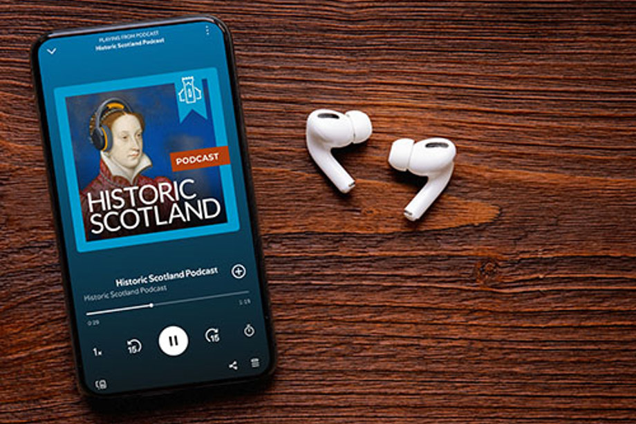 A set of ear buds and mobile phone on a wooden table showing an episode of the HS Members podcast 