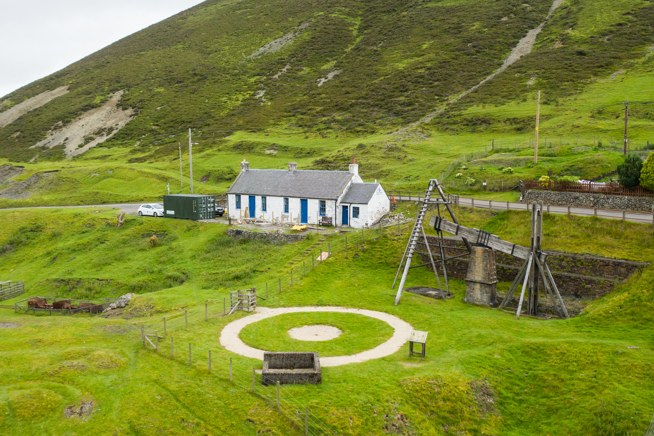A panoramic photo of a former lead mining landscape on a green but somewhat desolate hillside. It centres on a stone cottage and a wooden beam engine, which has a small circular track in front of it for horses to walk around.