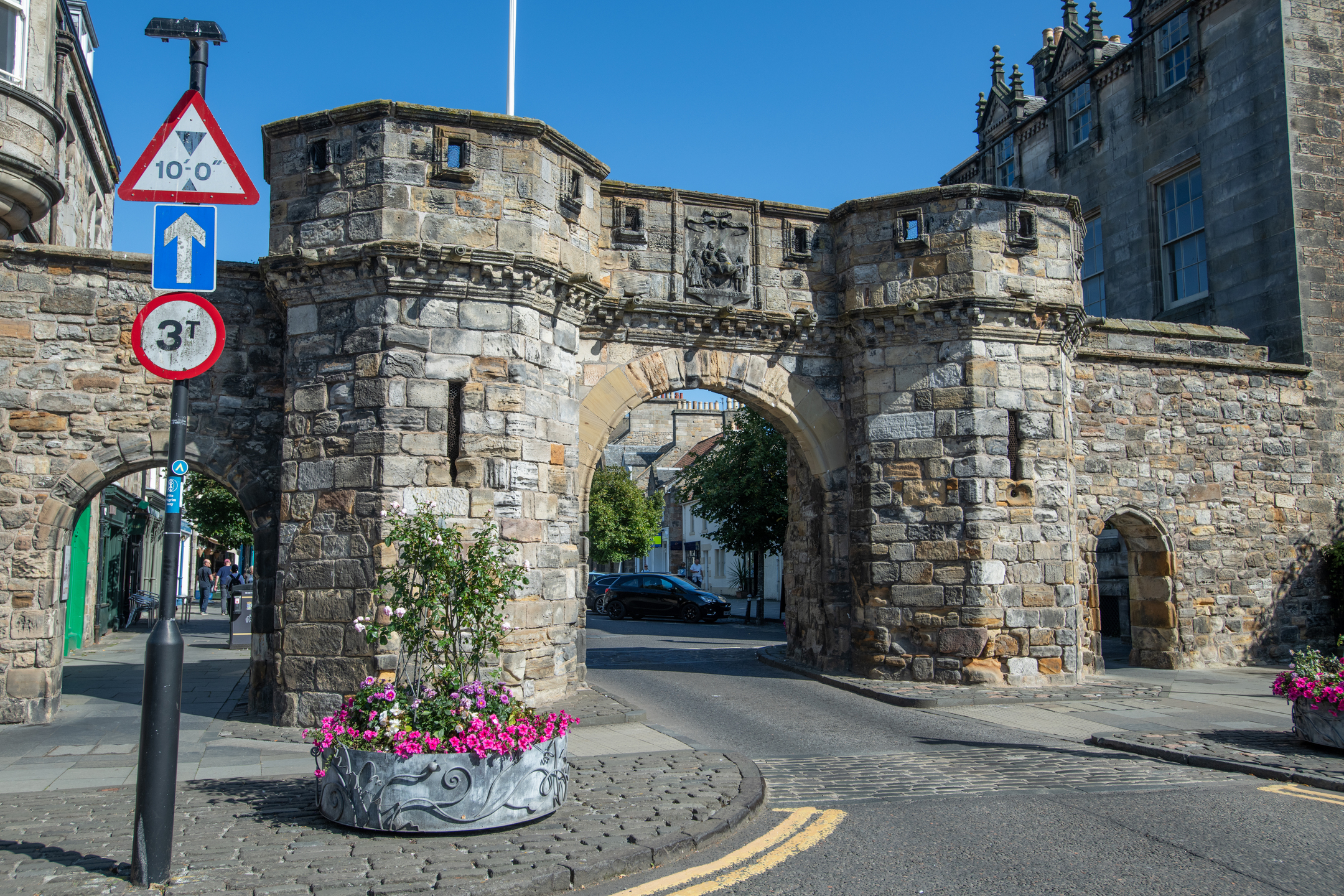 Close oblique view of the West Port gateway, showing the textured stone surface of the cylindrical tower on the left side. A triangular height‑restriction sign and a planter filled with pink flowers stand in front of the structure. Behind the gate, the central arch opens onto a narrow street lined with historic buildings.