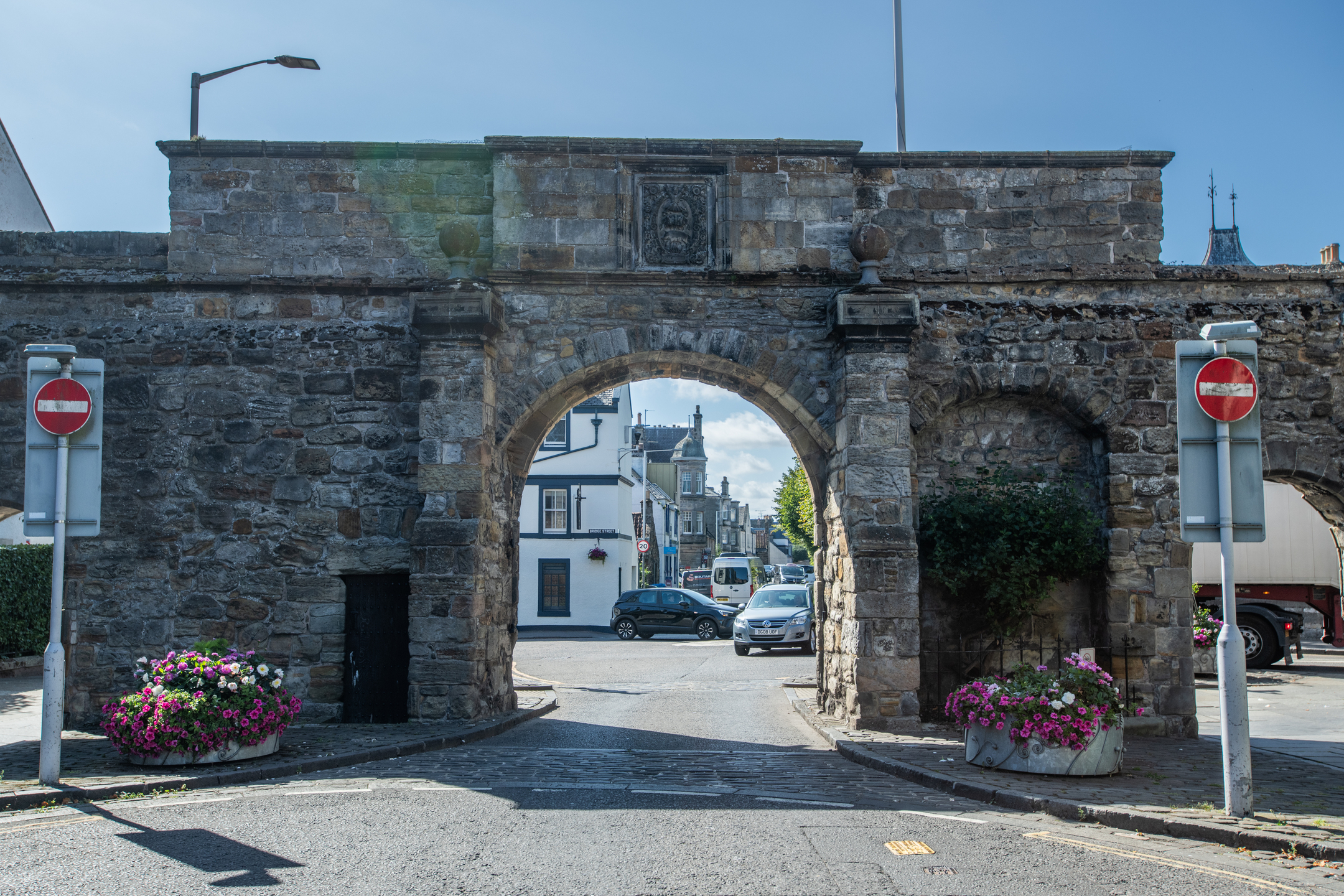 Front view of the West Port stone gateway showing its large central arch framed by two rounded towers made of weathered grey and brown stone. Through the arch, modern buildings, cars, and a sunlit street are visible. Two ‘no entry’ road signs and planters filled with pink flowers sit on either side of the structure.