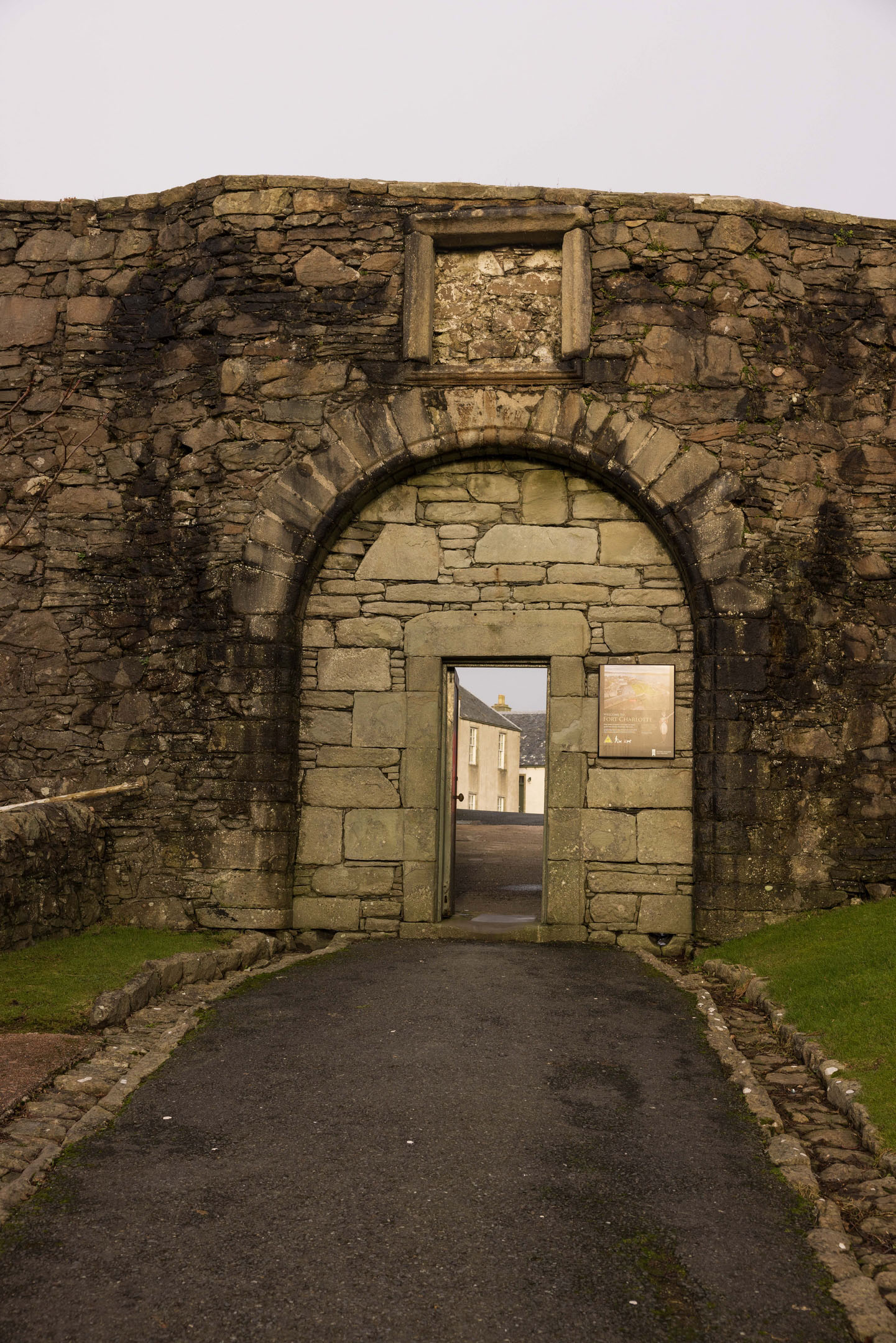 Tall stone archway with gate opening onto street and houses beyond.