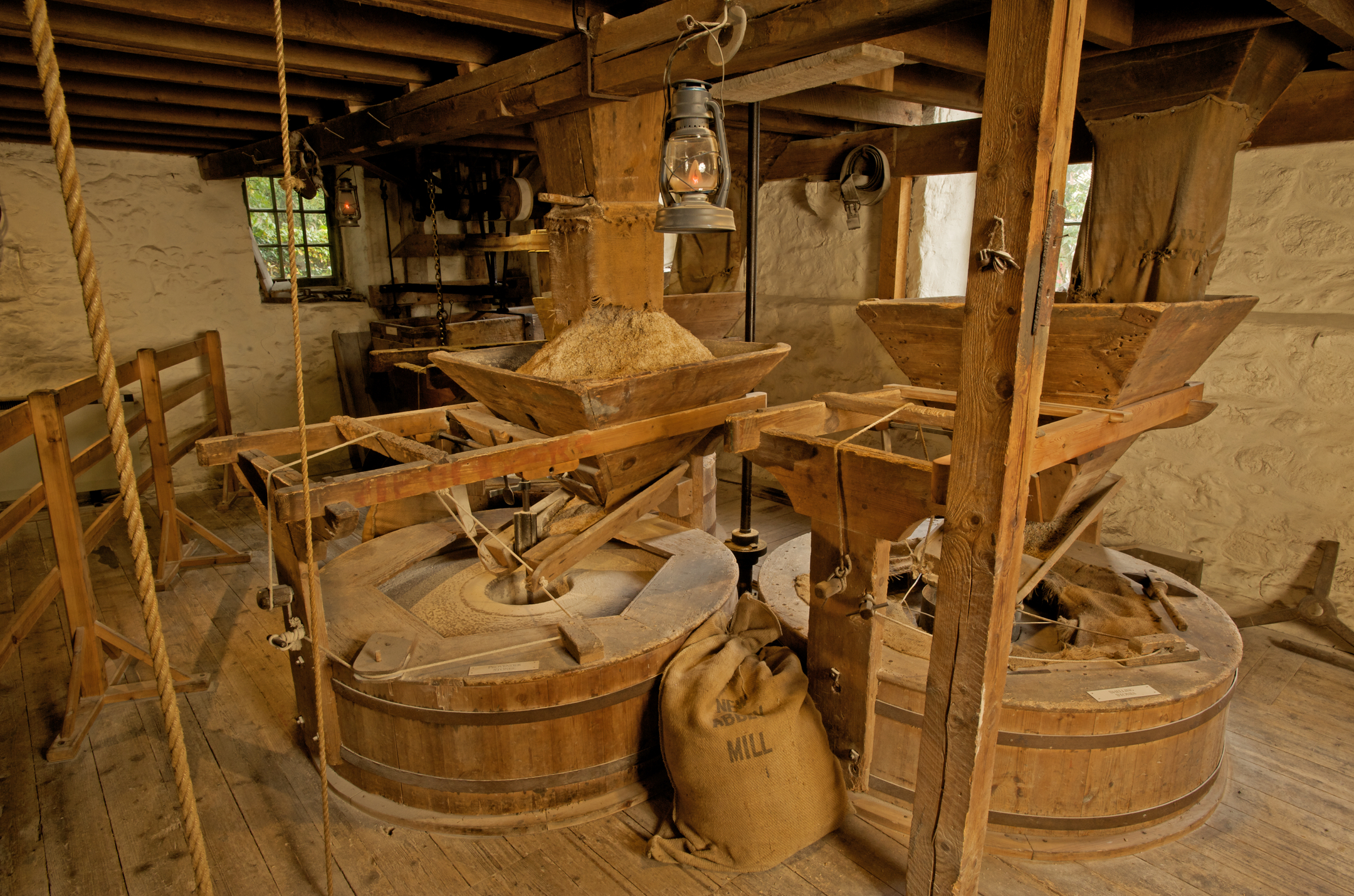 A wide view of the working interior of the mill, featuring two large circular millstones with wooden hoppers above them, ropes and beams overhead, and a sack labelled “New Abbey Mill” resting on the floor.