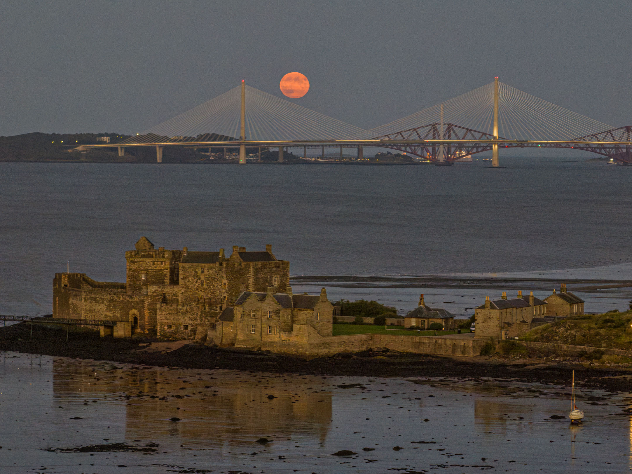 Blackness Castle on shoreline with bridges and rising orange moon in background.