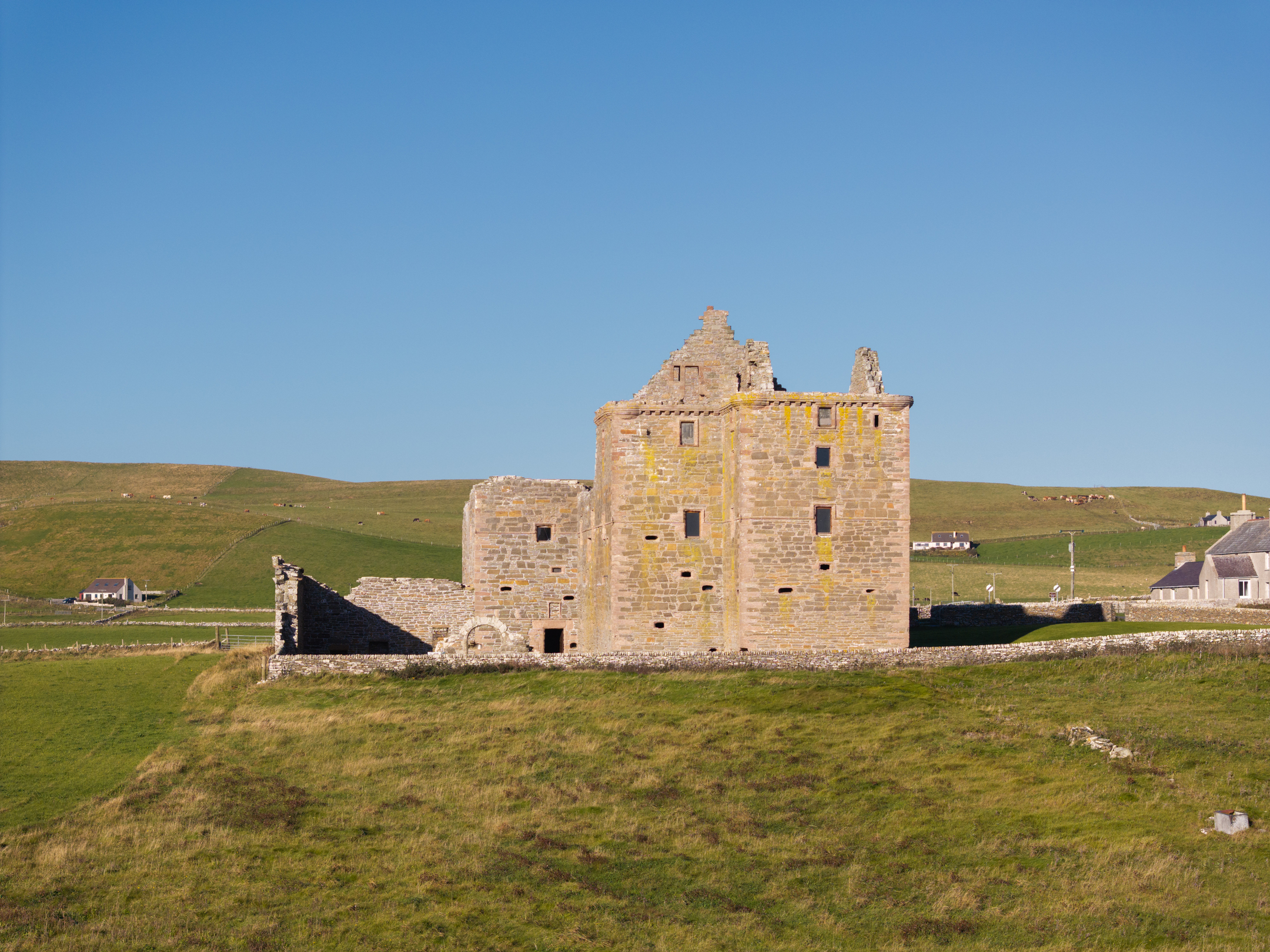 Wide view of Noltland Castle standing alone on open grassland, its tall stone walls illuminated by sunlight under a cloudless blue sky.