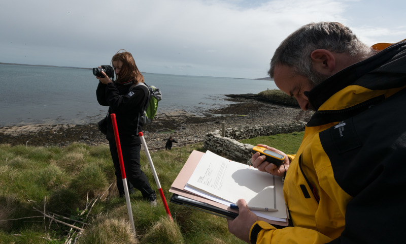 Two staff using equipment to record archaeology near a beach, one is looking a notes, the other is taking photos with a digital camera.
