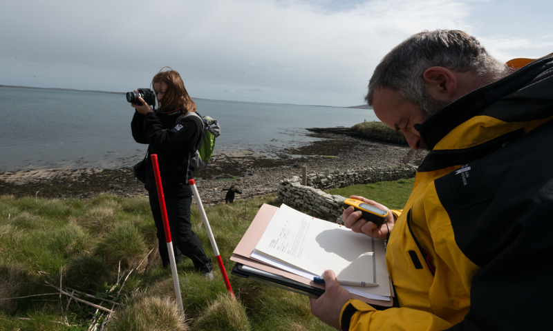 Two staff using equipment to record archaeology near a beach, one is looking a notes, the other is taking photos with a digital camera.