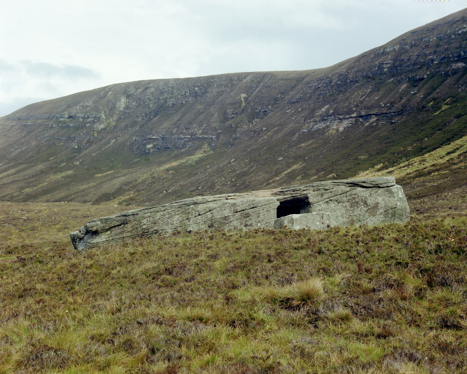 A colour photograph showing the Dwarfie Stane, a large rectangular sandstone boulder with a cut entrance, lying on open moorland. Rolling brown and green hills rise behind it under a cloudy sky.
