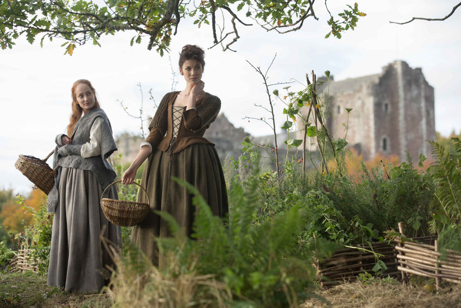 Two cast members from Outlander in a field with Doune Castle behind them