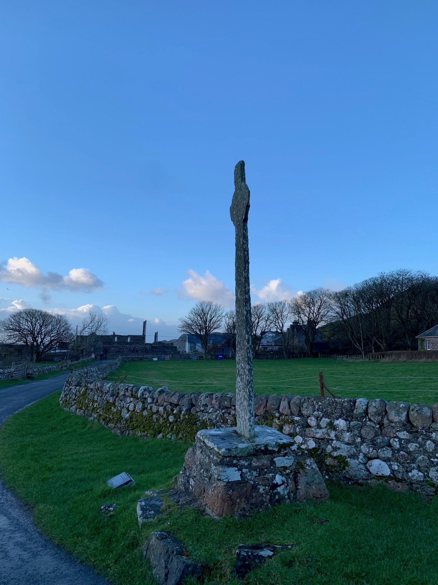 A tall, weathered stone cross mounted on a rough stone plinth beside a curving pathway and low dry‑stone wall. The cross stands in a grassy open area with scattered buildings and leafless trees in the distance under a bright blue sky with soft clouds.