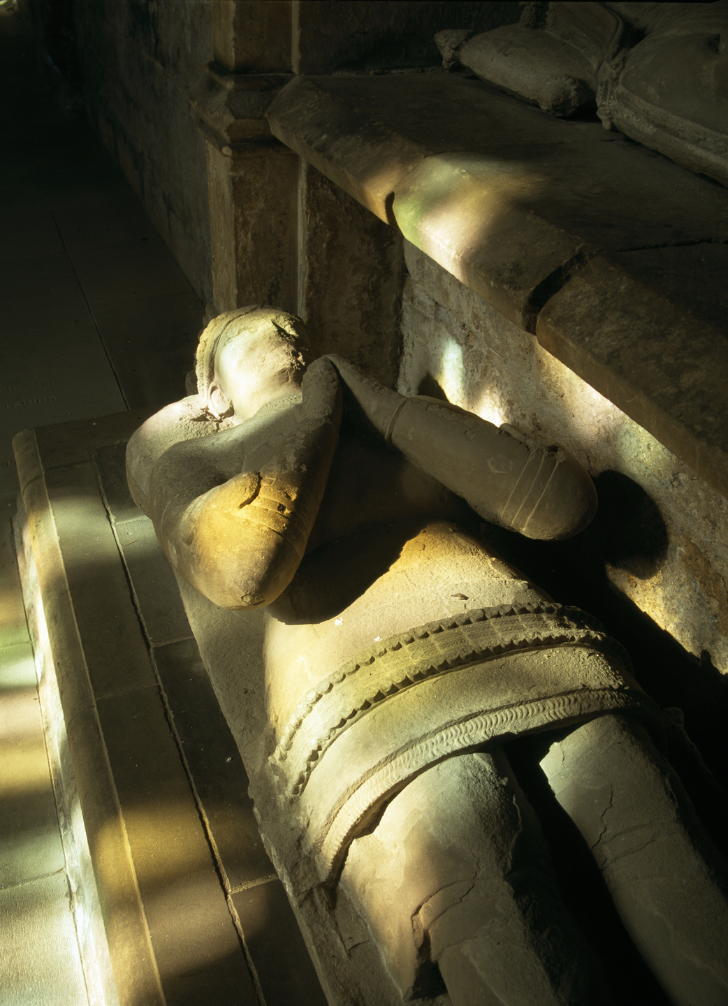 A stone effigy, laying down with hands clasped together facing up. There are colourful specks of light falling on it.