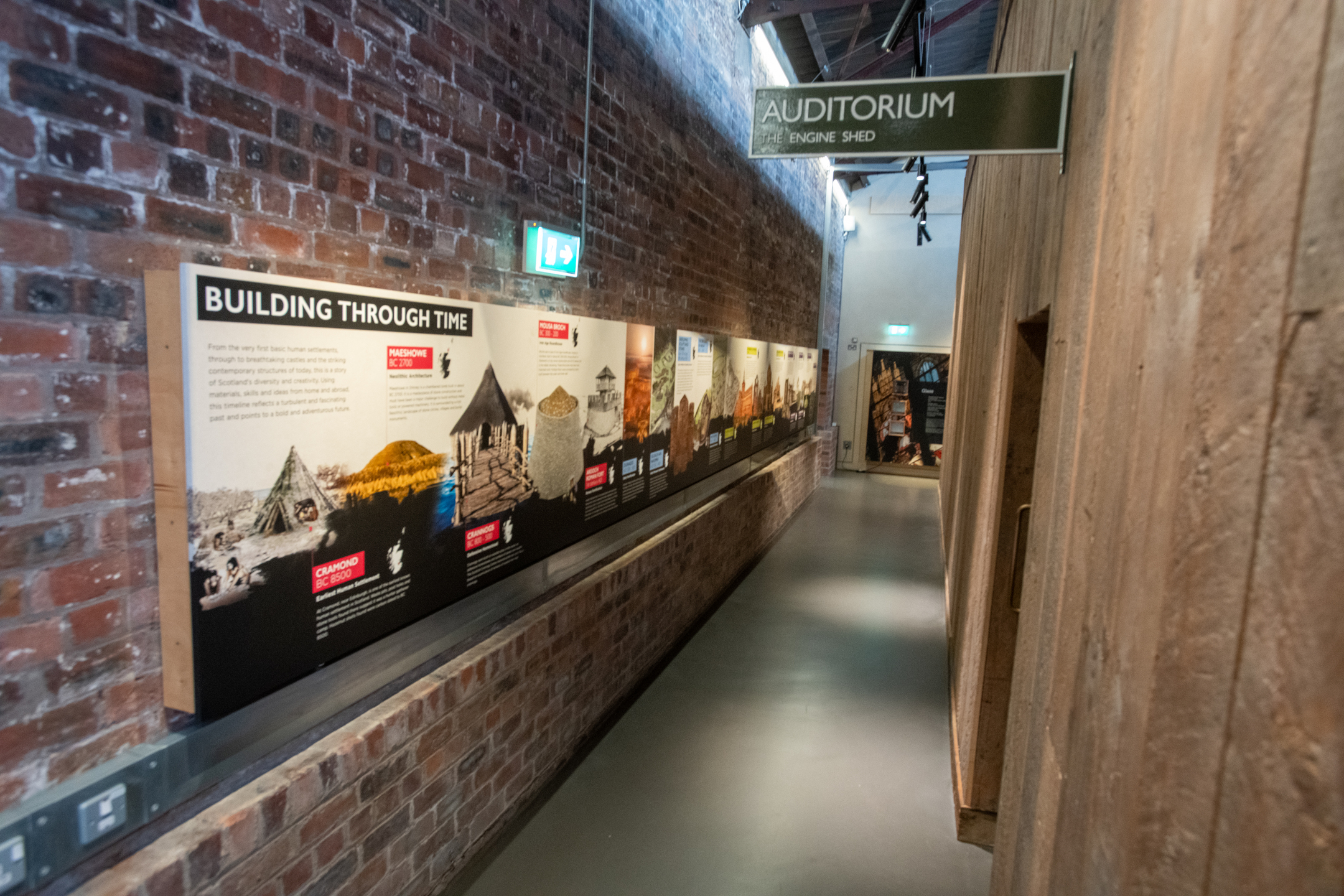 A corridor with wood panels and a sign that says 'auditorium' on the right, and a bare brick wall with a large interpretation panel showing a timeline on it.