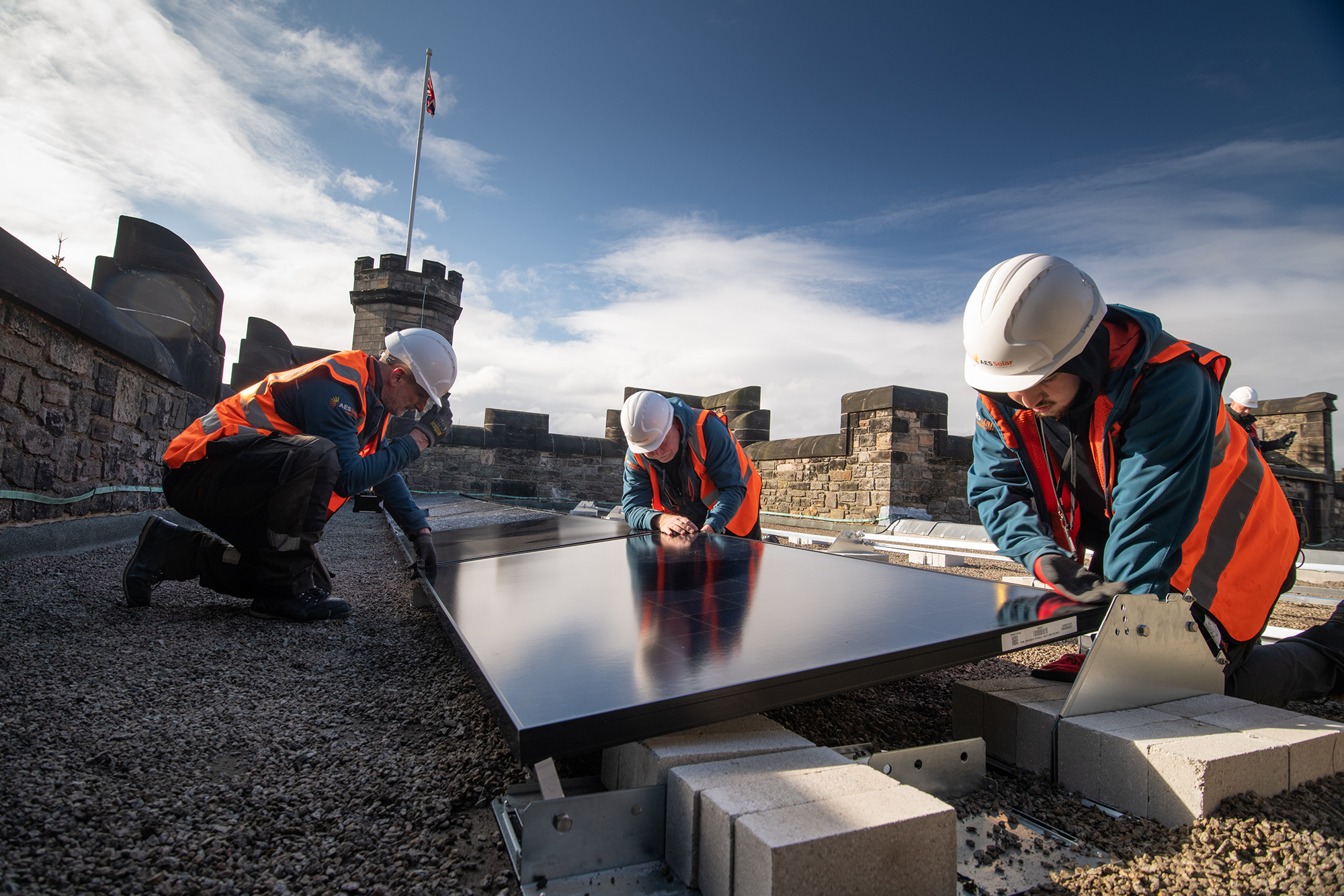 Three people in white hard hats and high-vis vests install a solar panel on a castle roof.