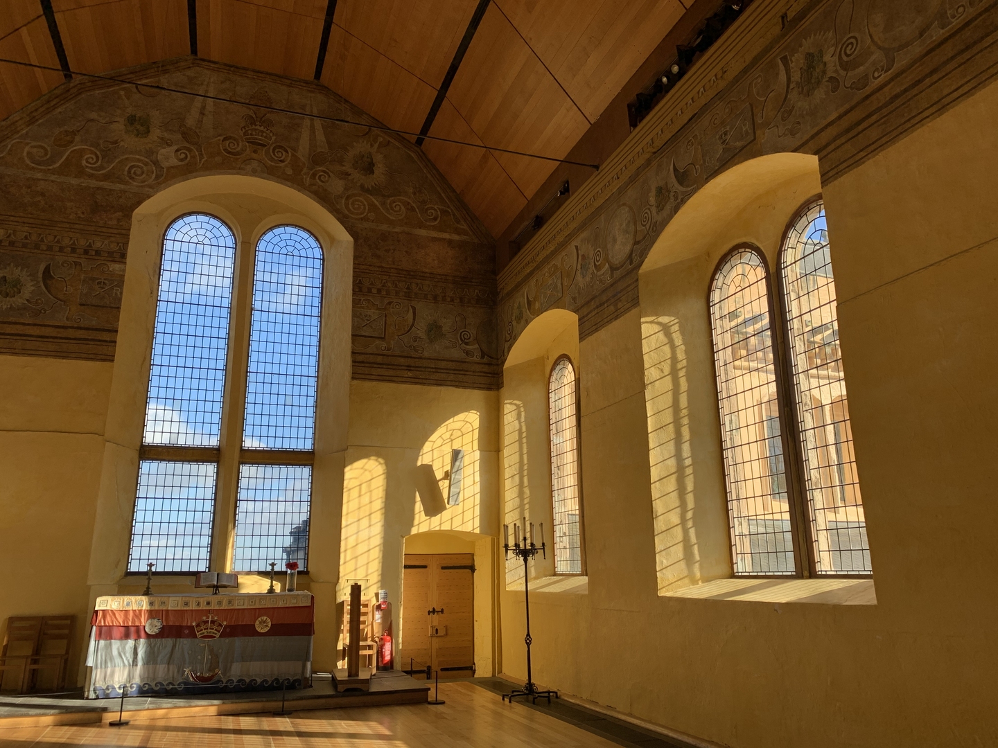 The corner of a chapel, with a high wooden ceiling, large arched windows and painted walls. There is light coming through the windows illuminating the room golden. There is a wide table at the front on a platform. 