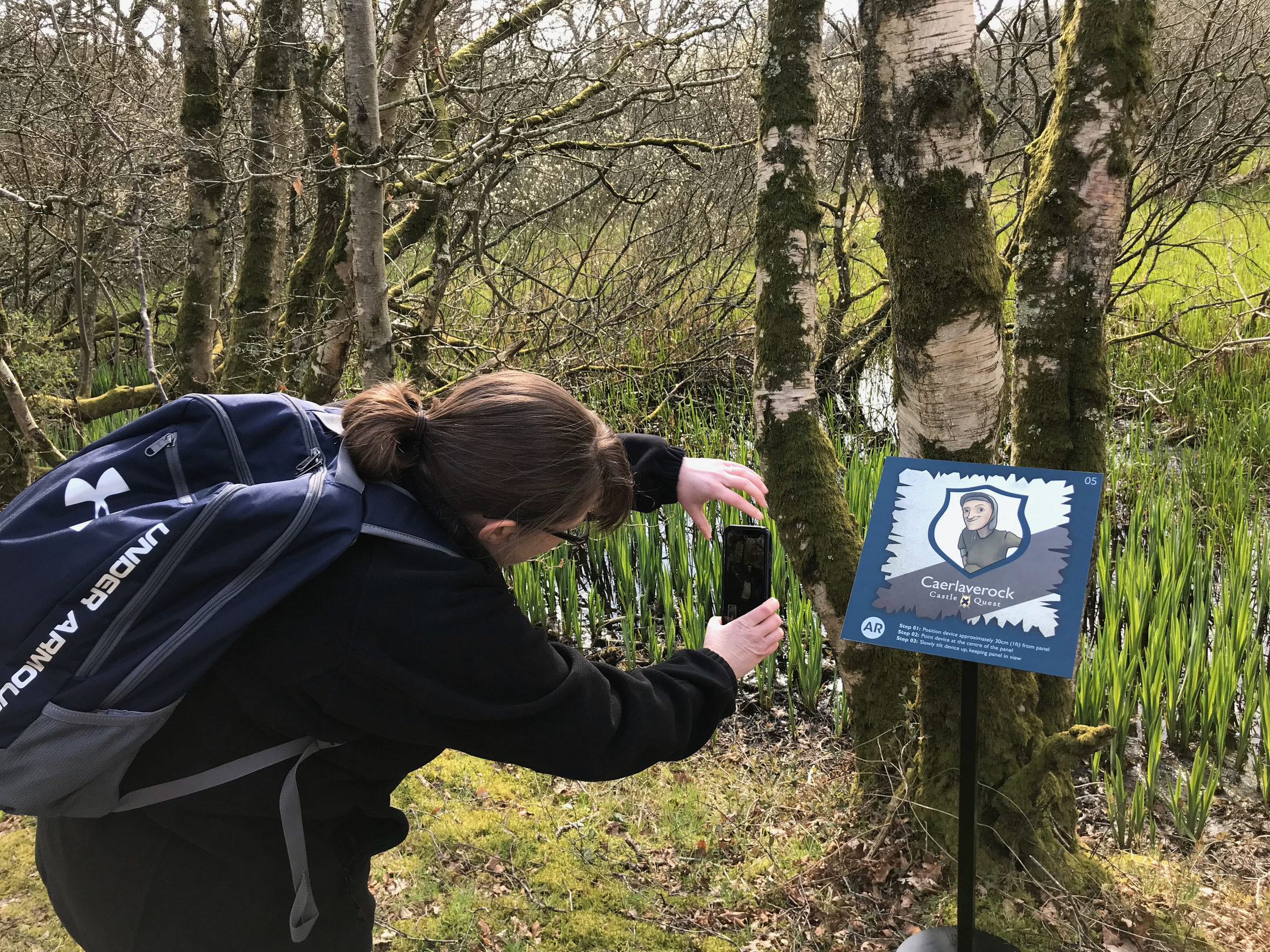 A visitor to Caerlaverock Castle uses their phone to scan a sign associated with the digital app available at the castle. The sign is located in woodland surrounding the ruin.