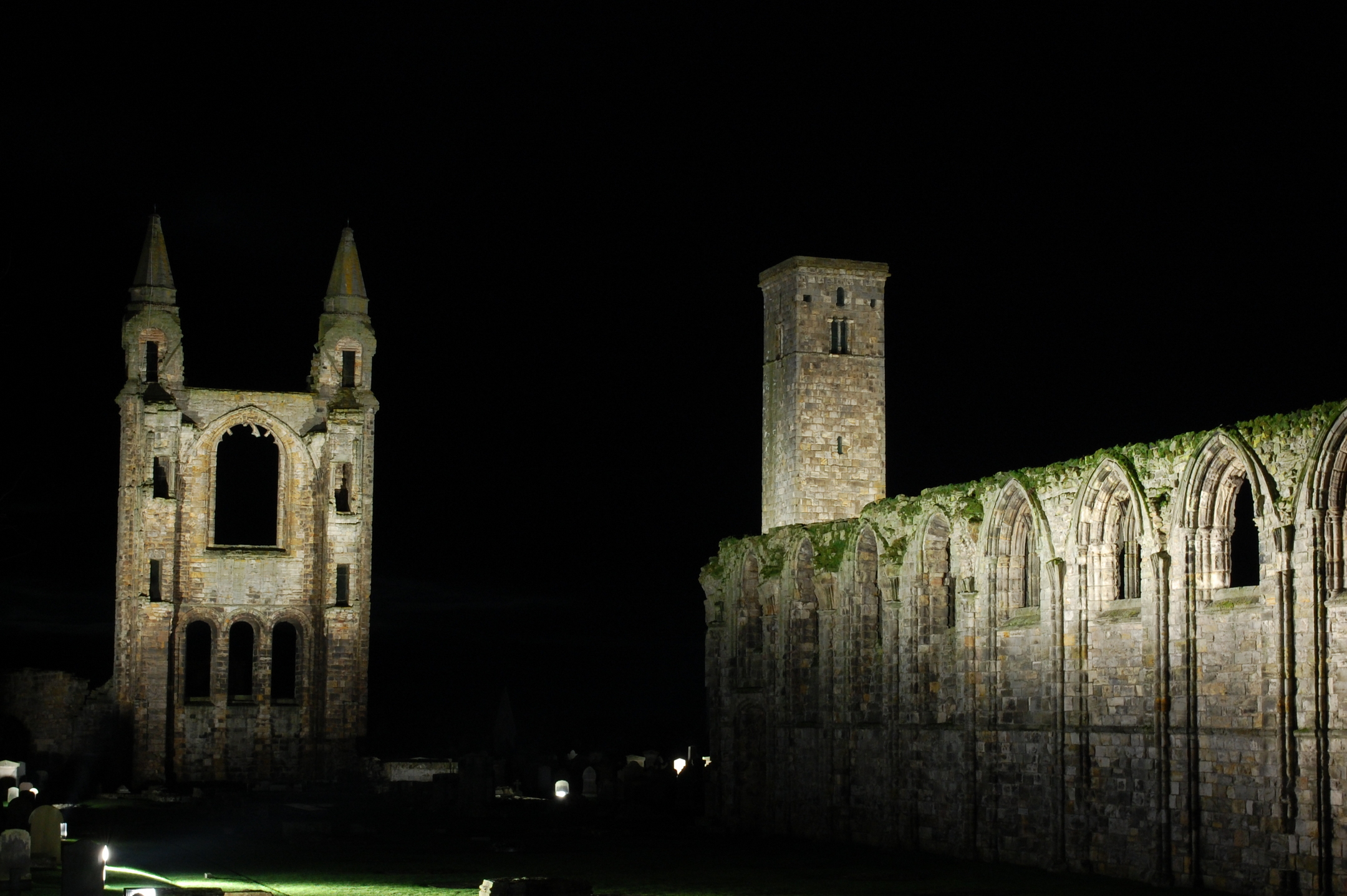 Floodlit stone ruins of a medieval cathedral stand against a completely dark night sky, with an illuminated tower and arched walls.