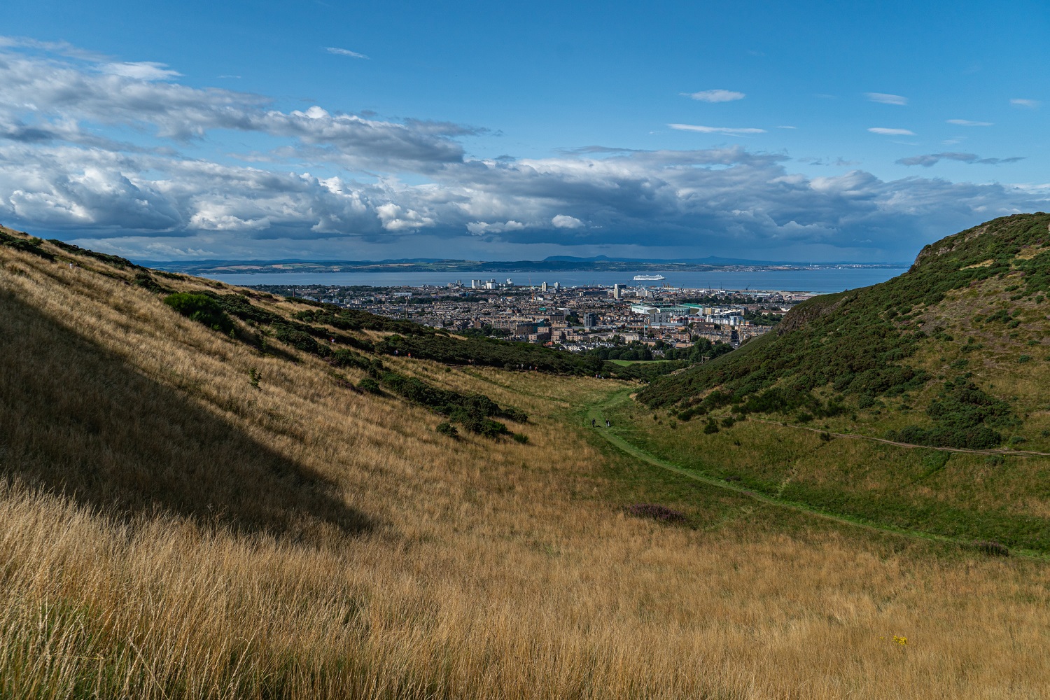 View of Hunter’s Bog in Holyrood Park, Edinburgh, showing a grassy valley with golden-brown slopes and green patches leading towards the city. Edinburgh’s skyline and the Firth of Forth are visible in the distance under a partly cloudy blue sky.