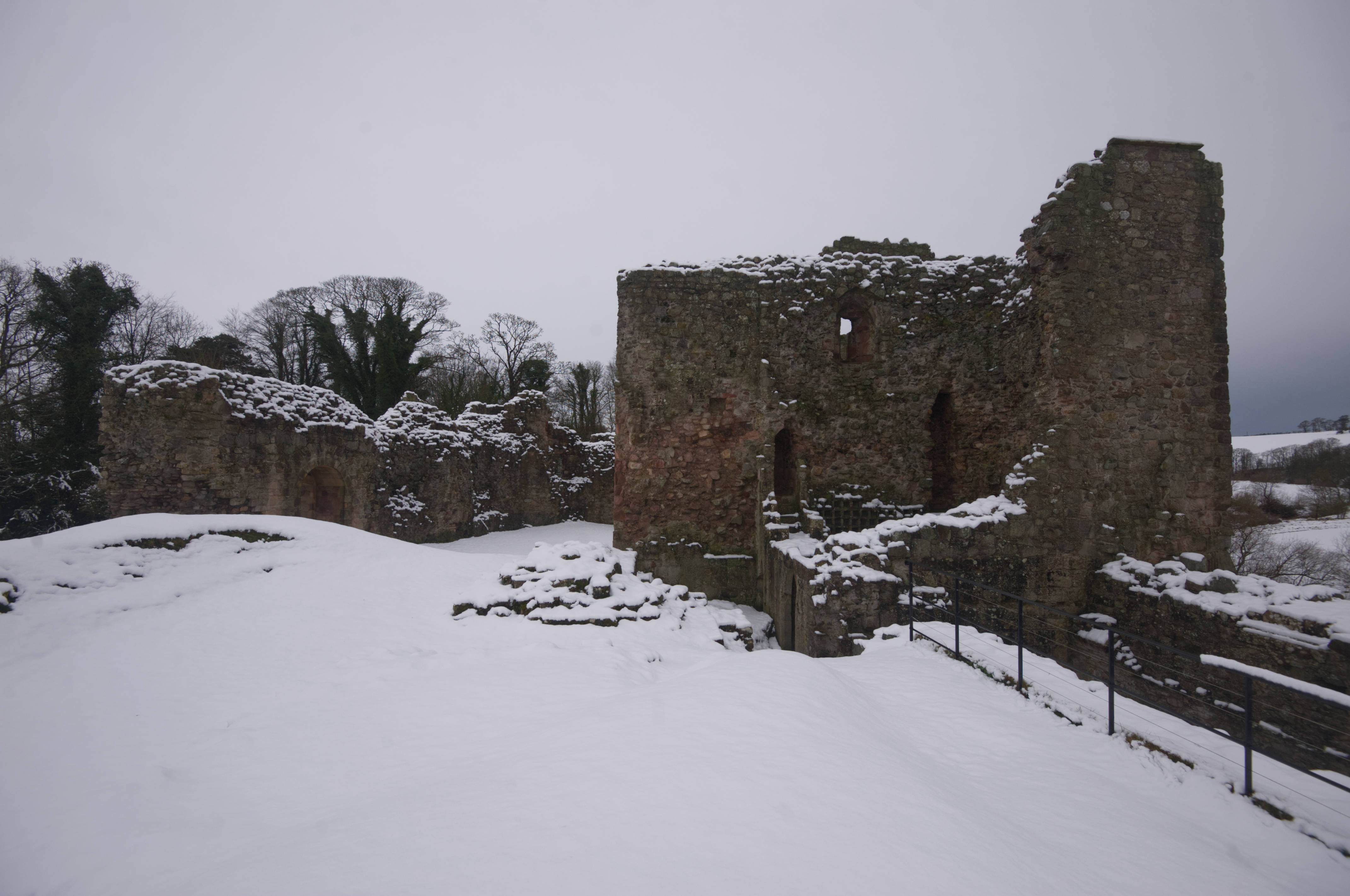 Ruins of Hailes Castle partially covered in snow, with bare trees behind and a grey winter sky overhead.