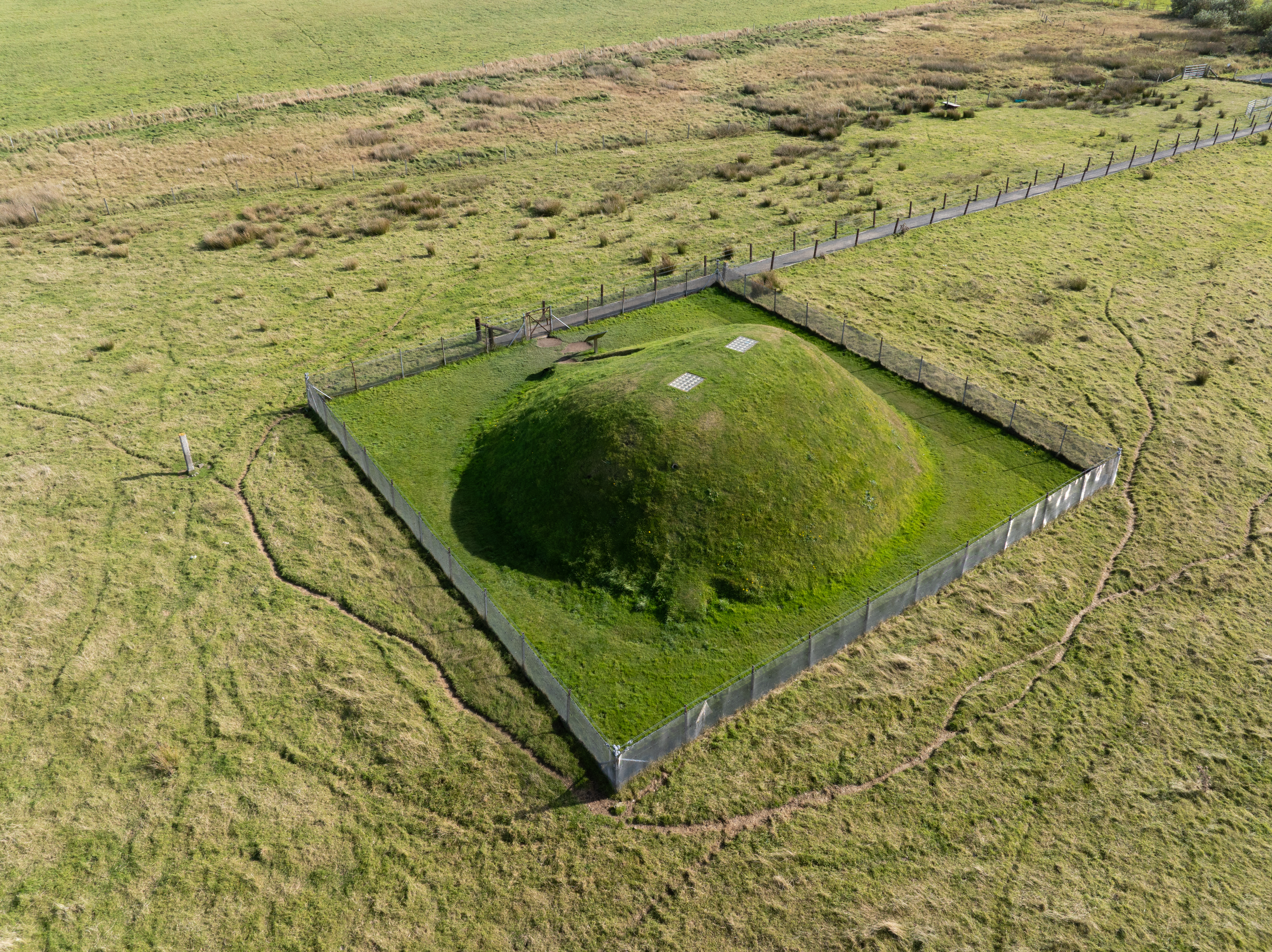 A large, grassy mound, fully fenced off, in the middle of a grassy field