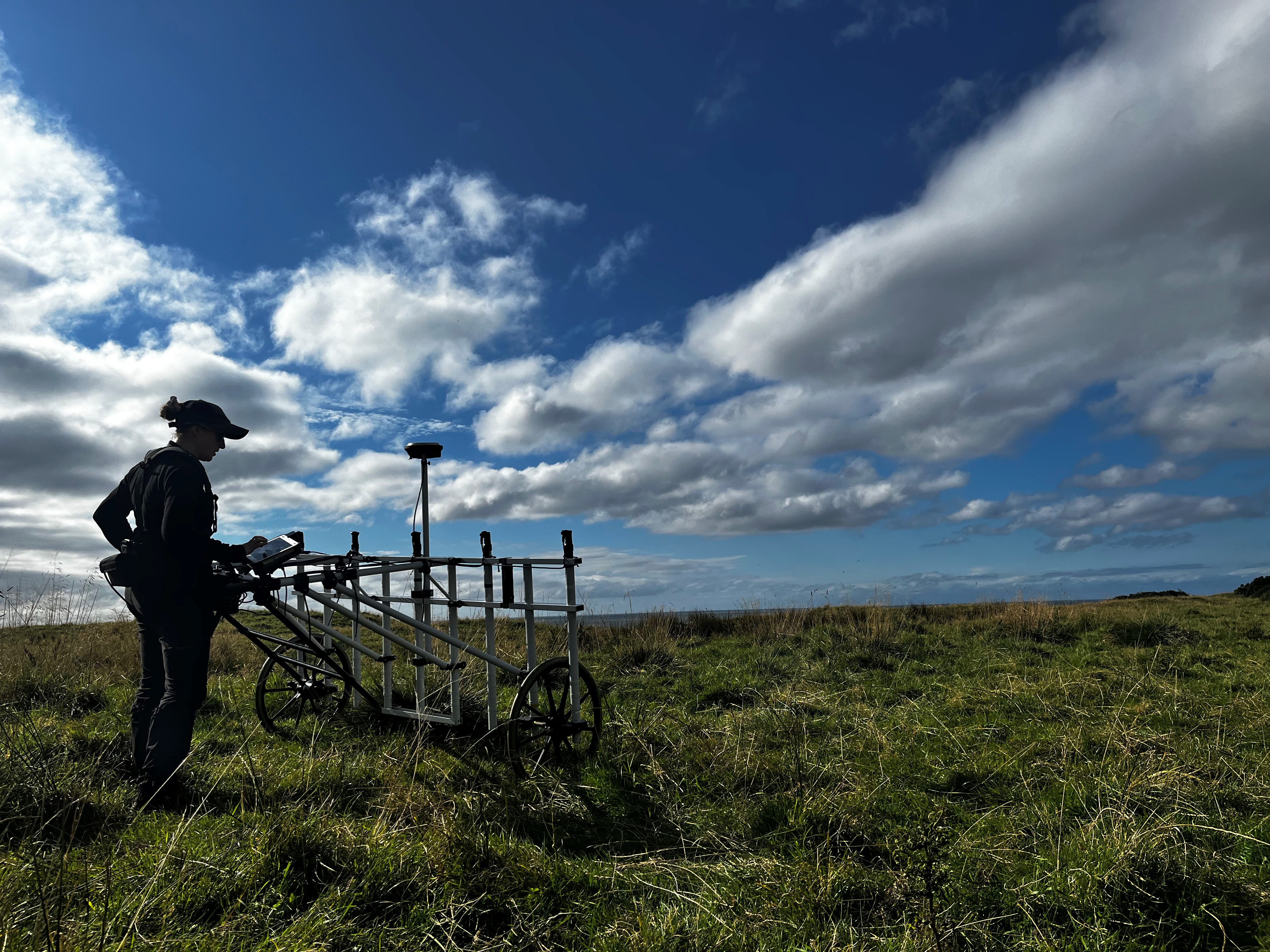  A silhouetted archaeologist operates a wheeled survey frame in windswept grassland beneath a wide blue sky filled with scattered clouds.