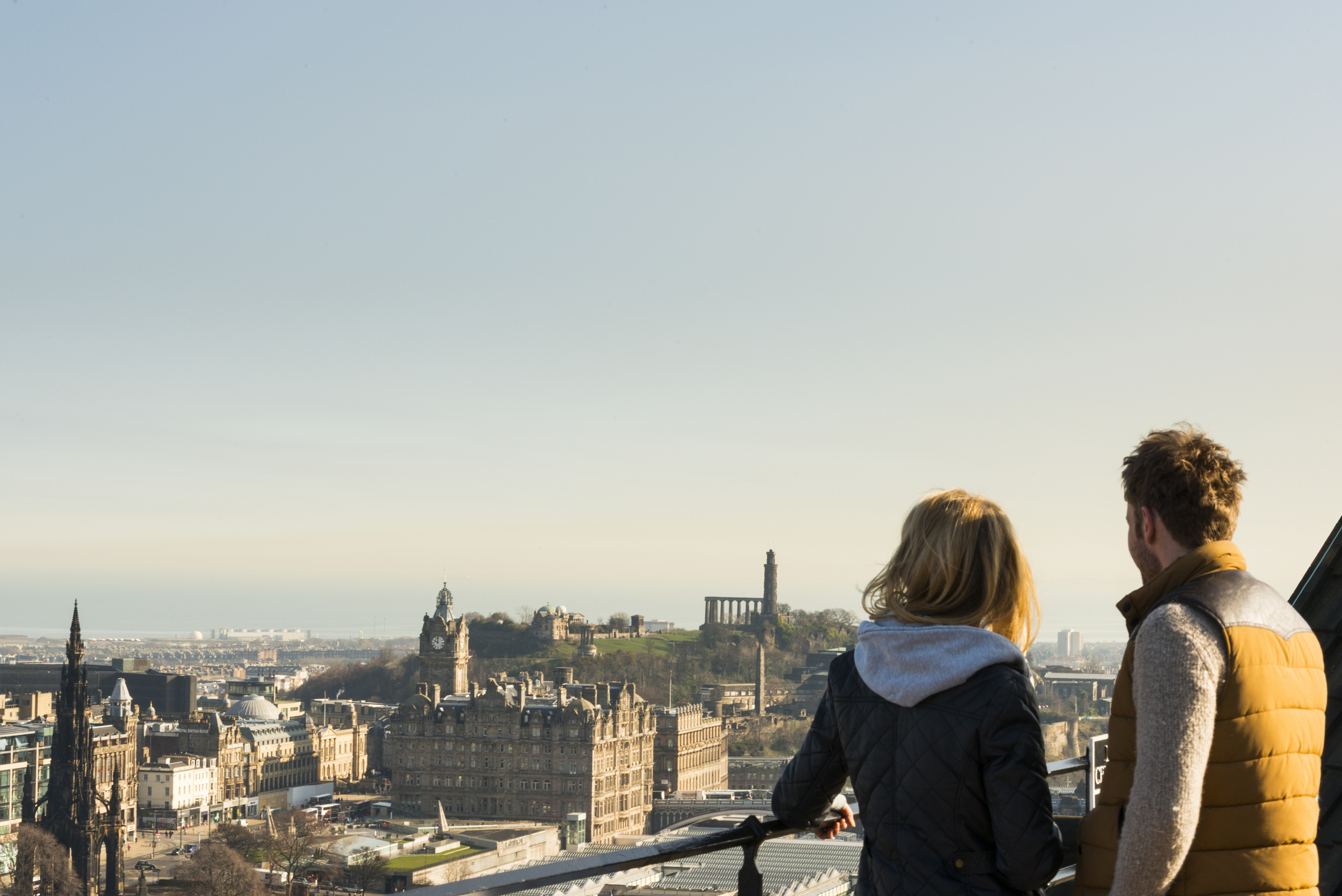 A man and a woman stood at a viewpoint looking over Edinburgh's skyline with the sea in the background