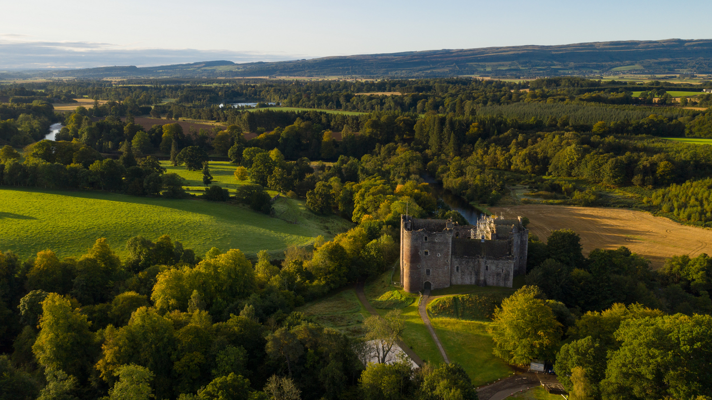 Aerial view of a historical stone castle, in a rural landscape. It sits on grass, with a path leading away from it. There are trees and forests in the background, as well as fields. 